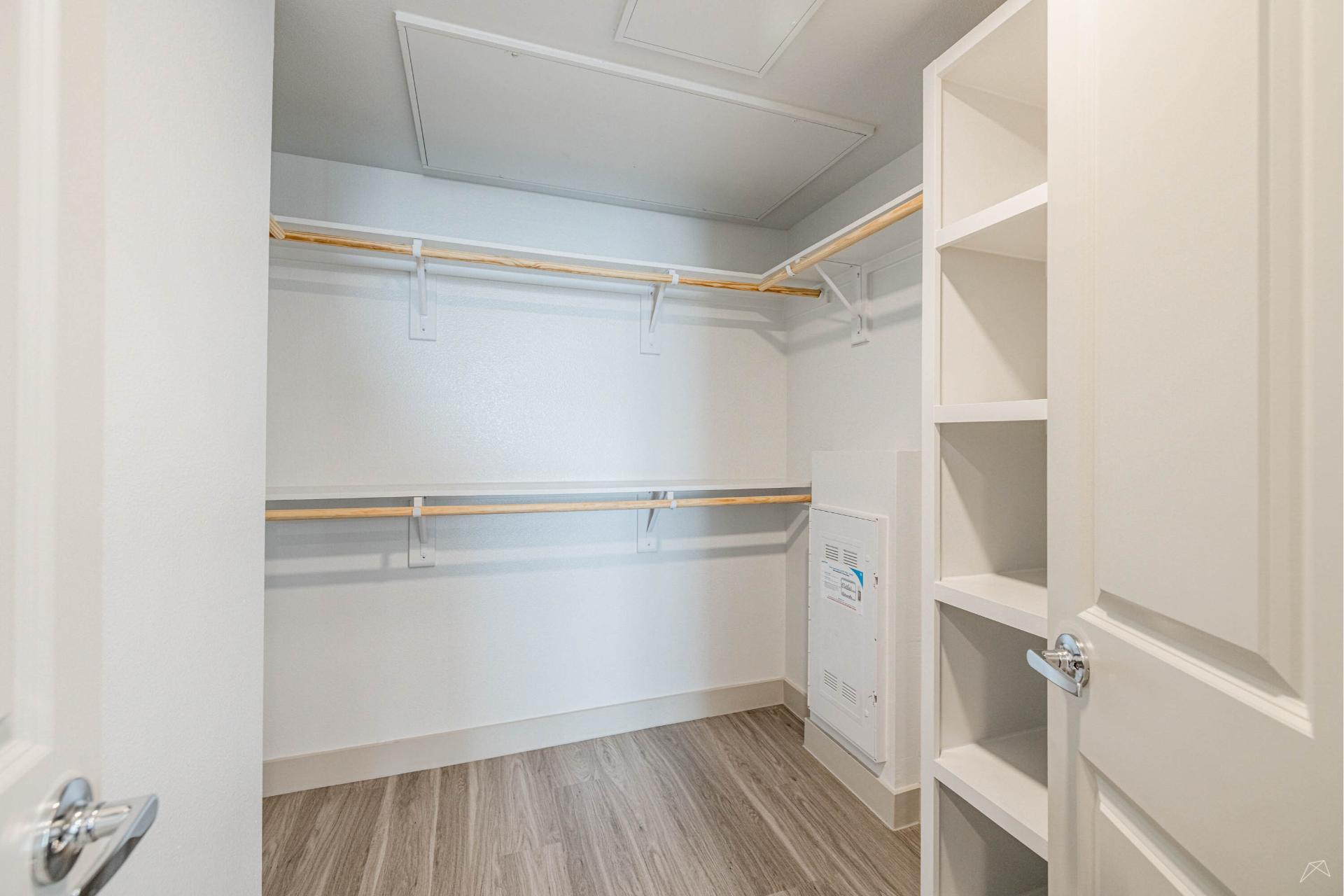 A spacious, empty walk-in closet with light wood-look flooring, white walls, built-in shelves right, hanging rods on two walls, open door.