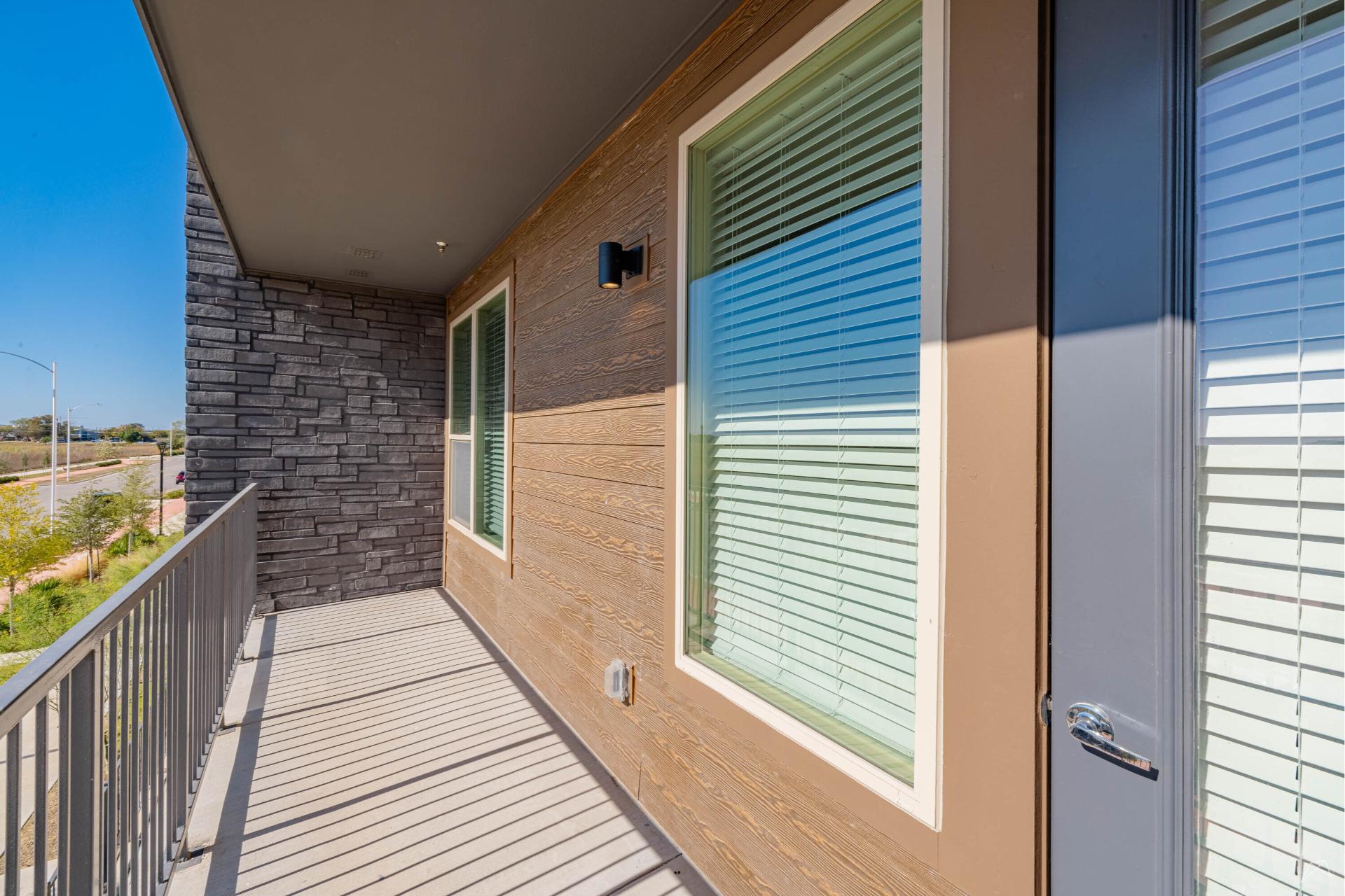 A modern apartment balcony with metal railing, stone and wood-style siding, two windows with white blinds, overlooking a sunny street and greenery.