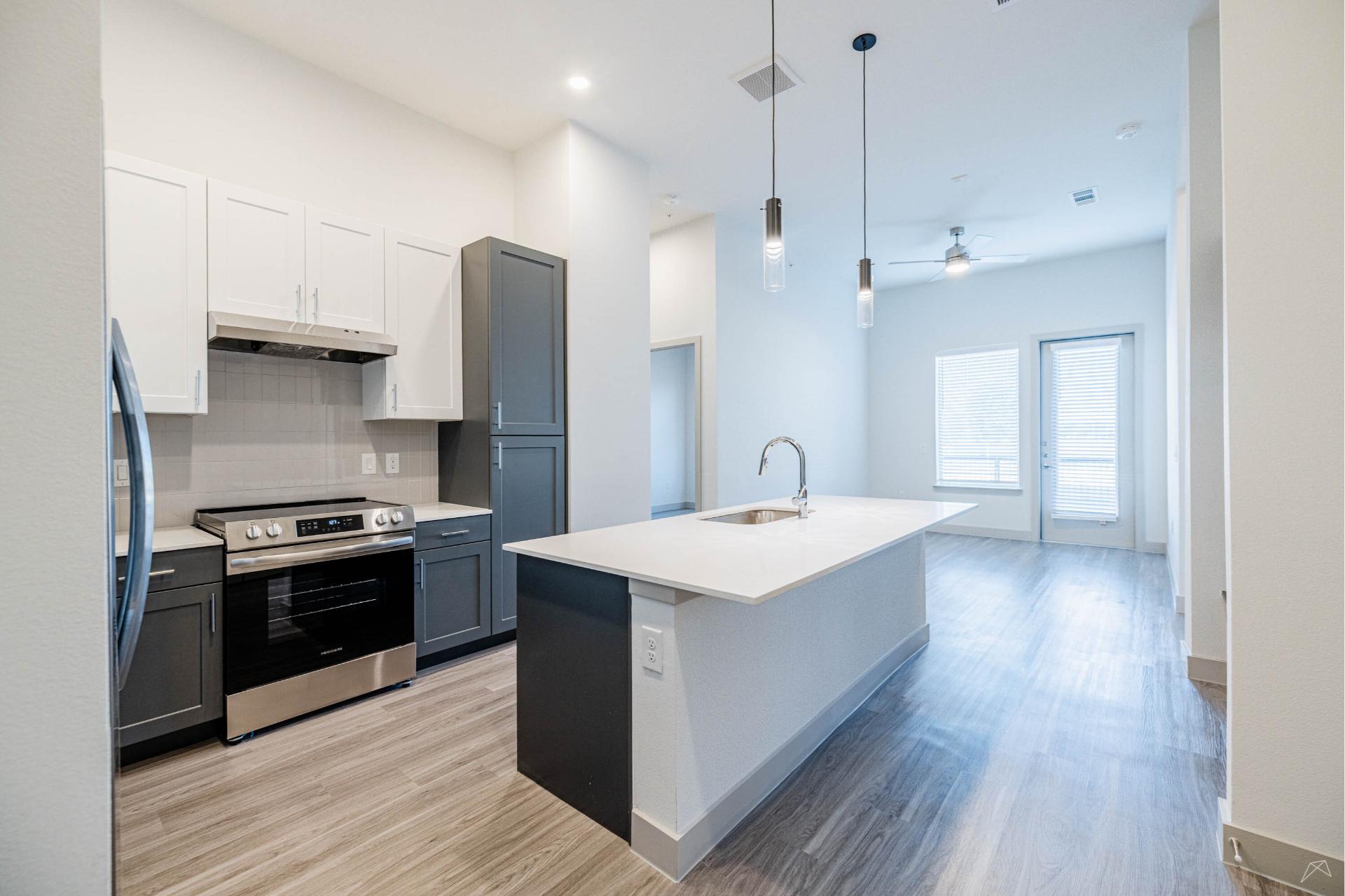 Modern kitchen with white and gray cabinets, stainless appliances, a large island with sink, pendant lights, wood-style flooring, and bright living area.