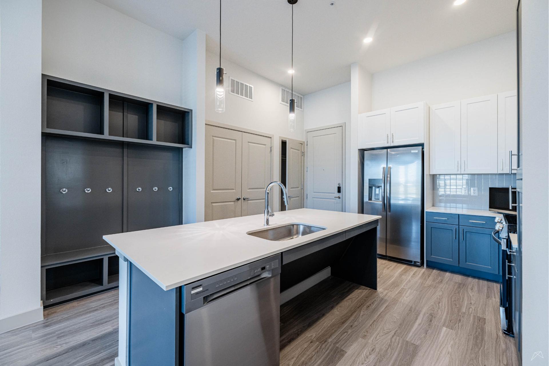 Modern kitchen with a large island, stainless steel appliances, blue and white cabinetry, pendant lights, wood-style flooring, and a coat rack with cubbies on the left.
