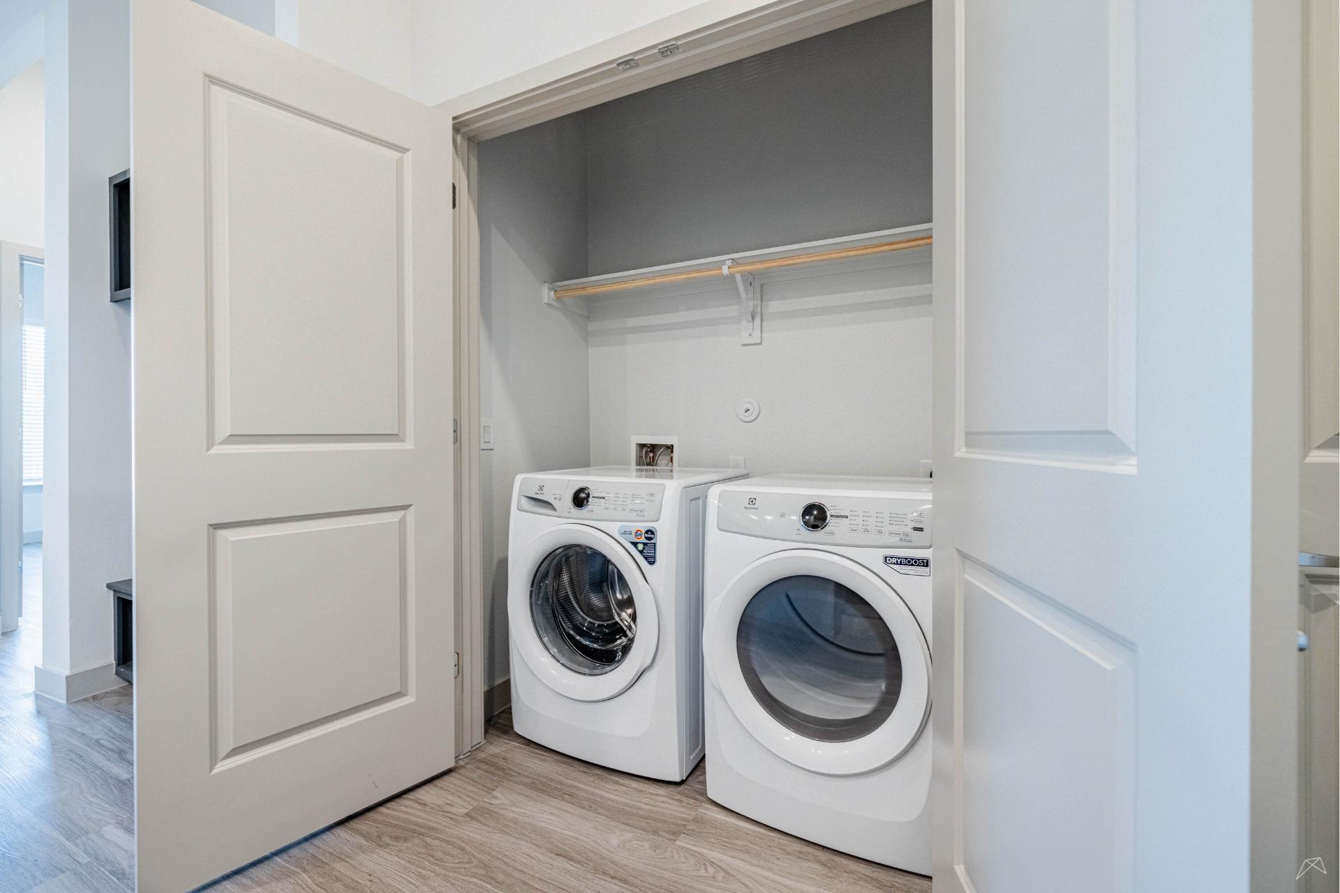 A modern laundry closet with white bi-fold doors, front-loading washer and dryer, beneath a shelf on a light wood-look floor.