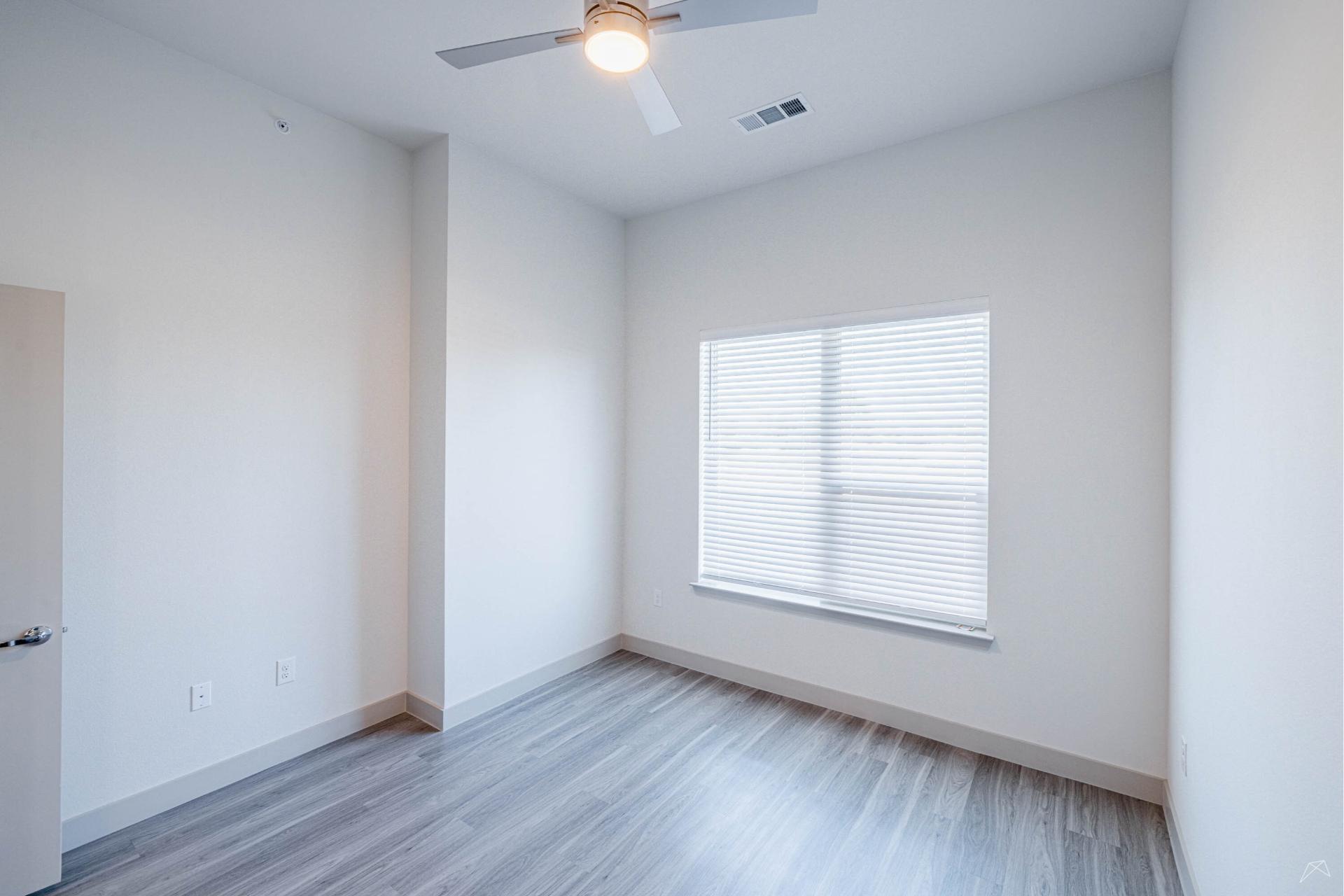 Bright, empty room with light gray wood-look flooring, white walls, a large window with closed blinds, and a ceiling fan with light.