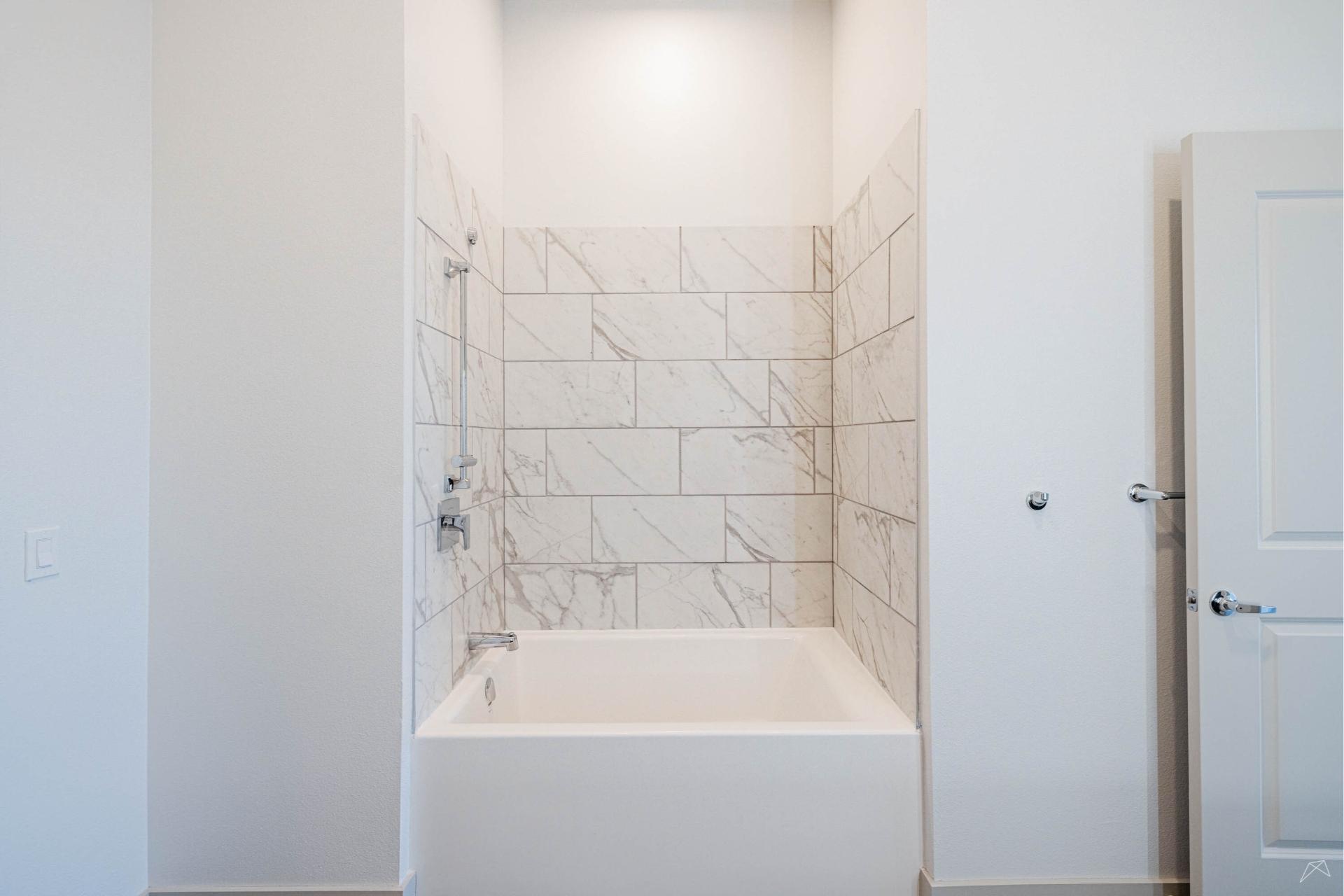 A modern white bathtub with marble-tiled walls and a silver showerhead in a bright, minimal bathroom and a closed white door to the right.