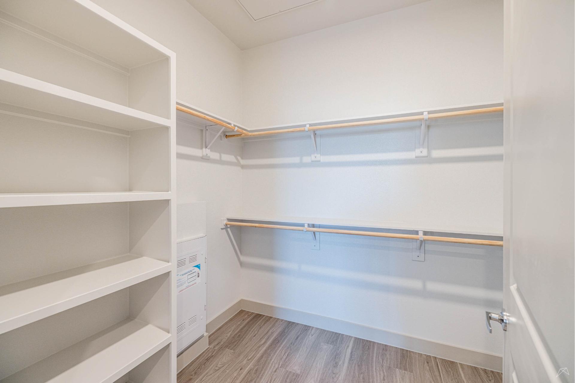 A walk-in closet with white walls, built-in shelves on the left, two rows of wood-style hanging rods, and light wood-look flooring.