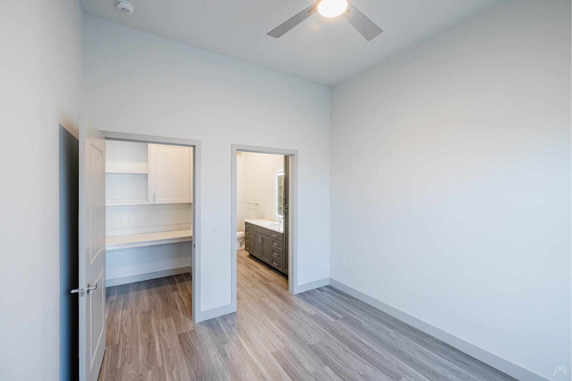 A bright, empty room with light gray walls, wood-look flooring, open doors to a small office nook and bath. Ceiling fan with light.