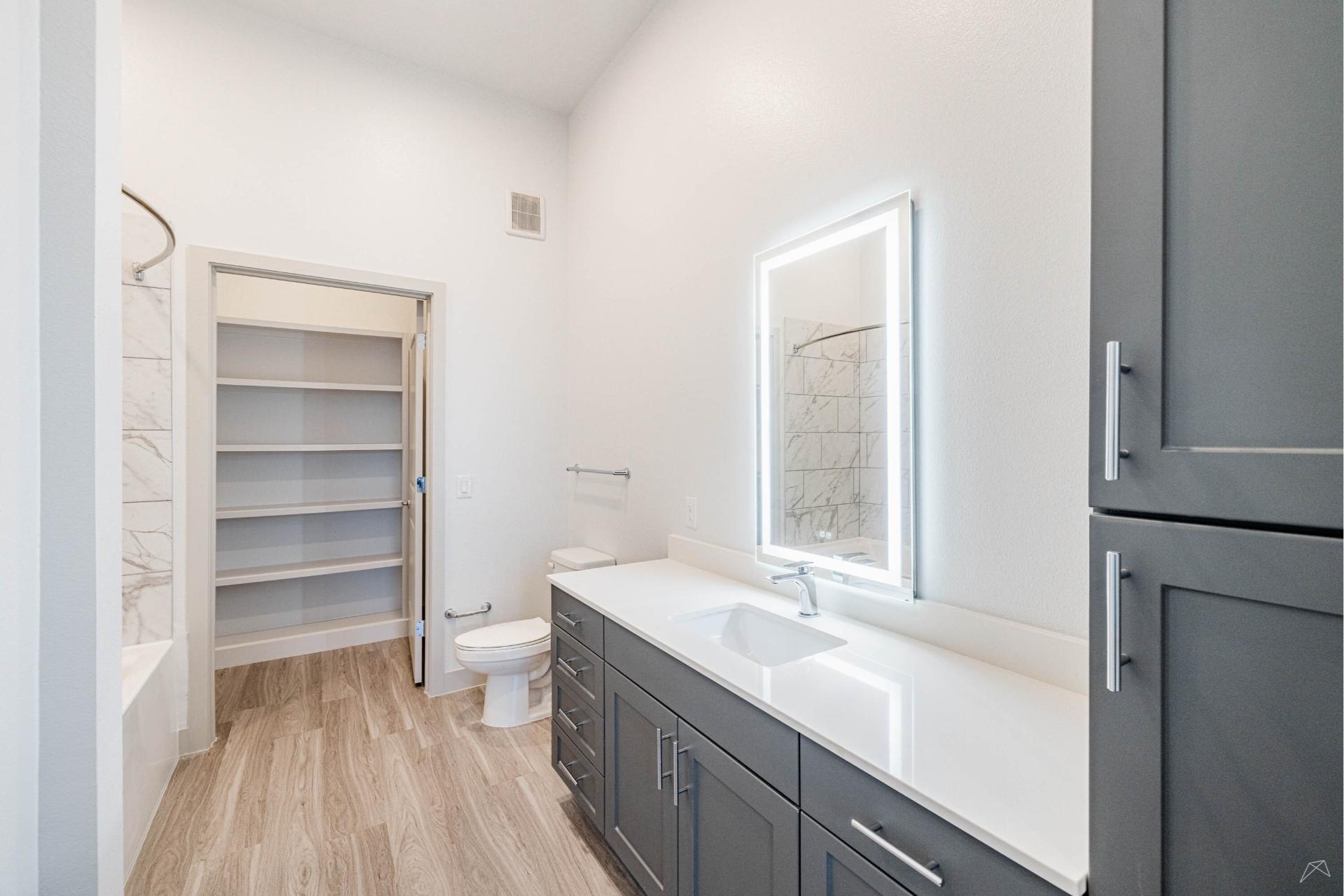 Modern bathroom with gray cabinets, large illuminated mirror, white countertops, toilet, and wood-look flooring for a bright feel.