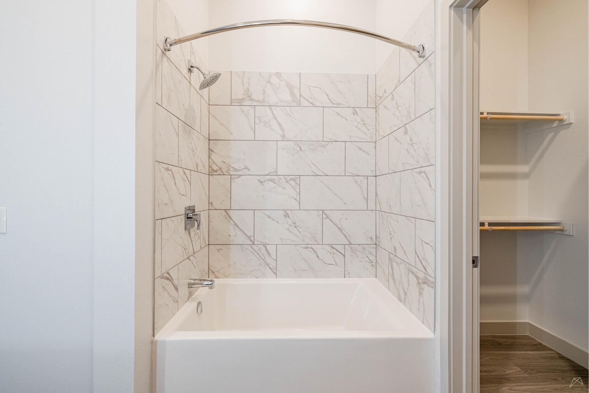 A modern bathroom with a white bathtub, marble-patterned tile walls, a curved shower rod, and an open closet with wood-look shelves.