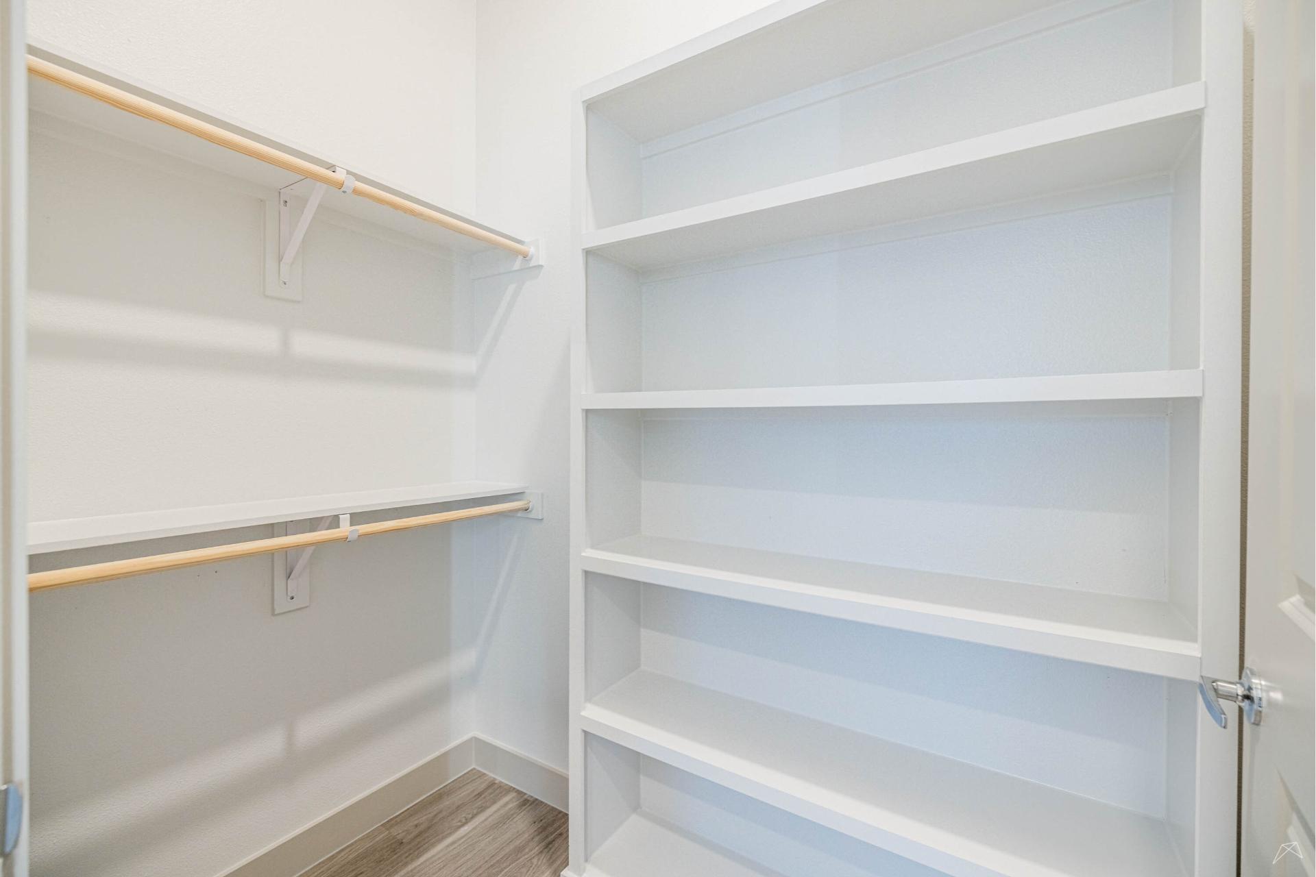 A clean, empty walk-in closet with white shelves on the right and two wood-style hanging rods with shelves on the left. Light wood-look floor and white walls.