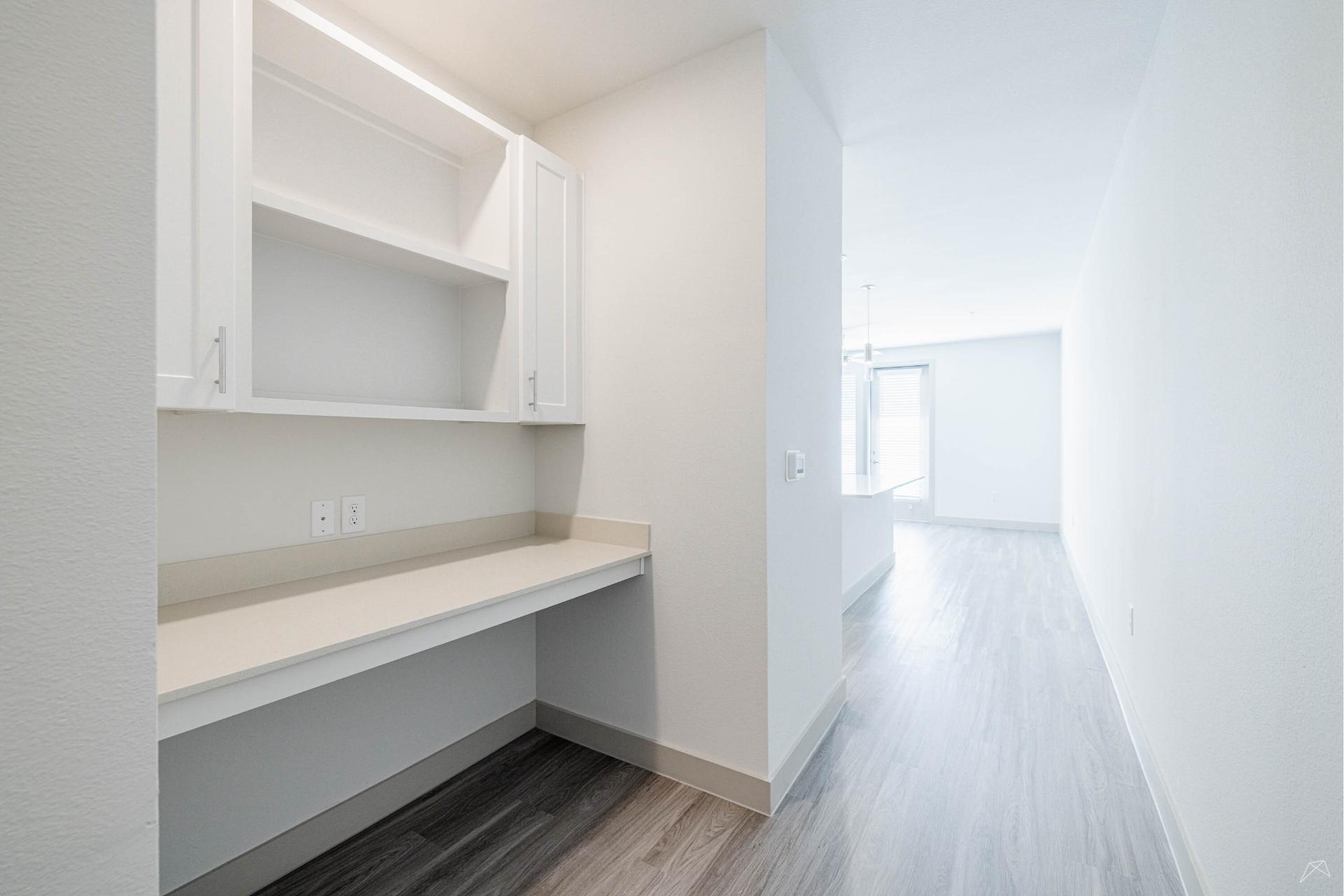 A small built-in desk with open shelves and cabinets sits by a white wall, light countertops, wood-look flooring, and big bright windows.