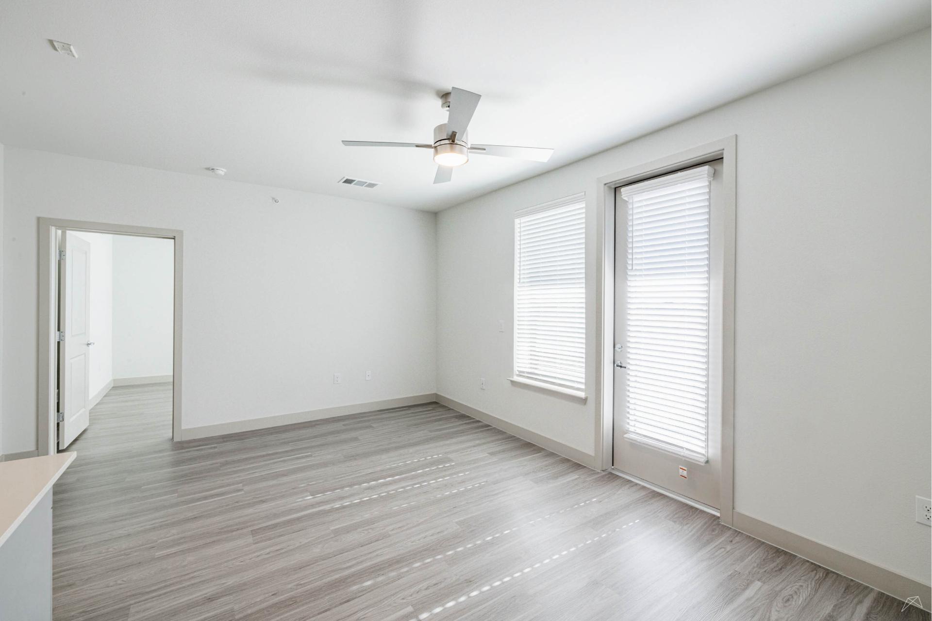A bright, empty room with light gray wood-look flooring, white walls, two large windows with blinds, a glass door, and a ceiling fan. An open doorway leads to another room.