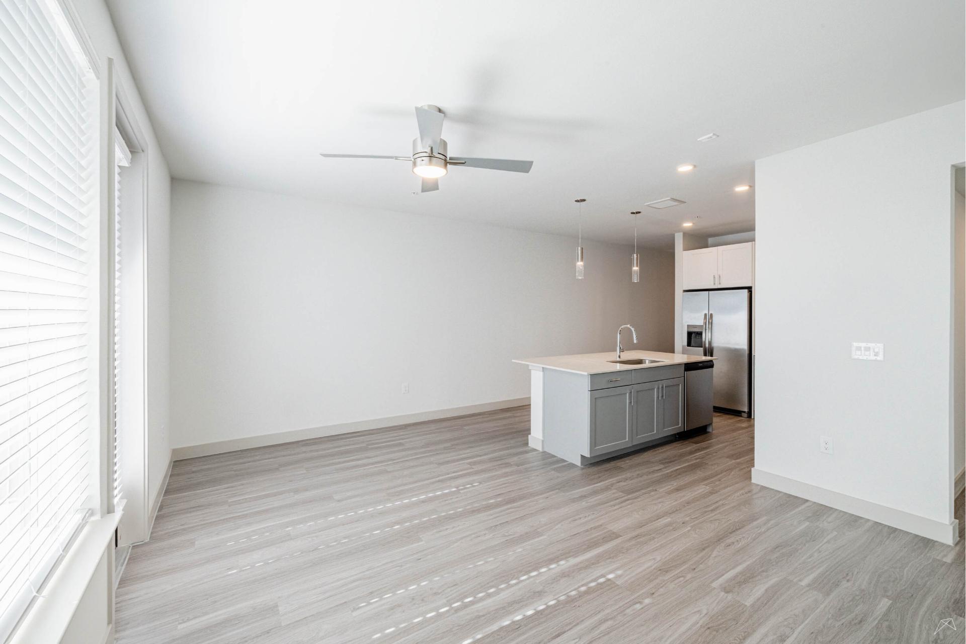 Bright, modern apartment with light gray wood-style floors, large windows, an open kitchen island, stainless appliances, and pendant lights. Ceiling fan in main area.
