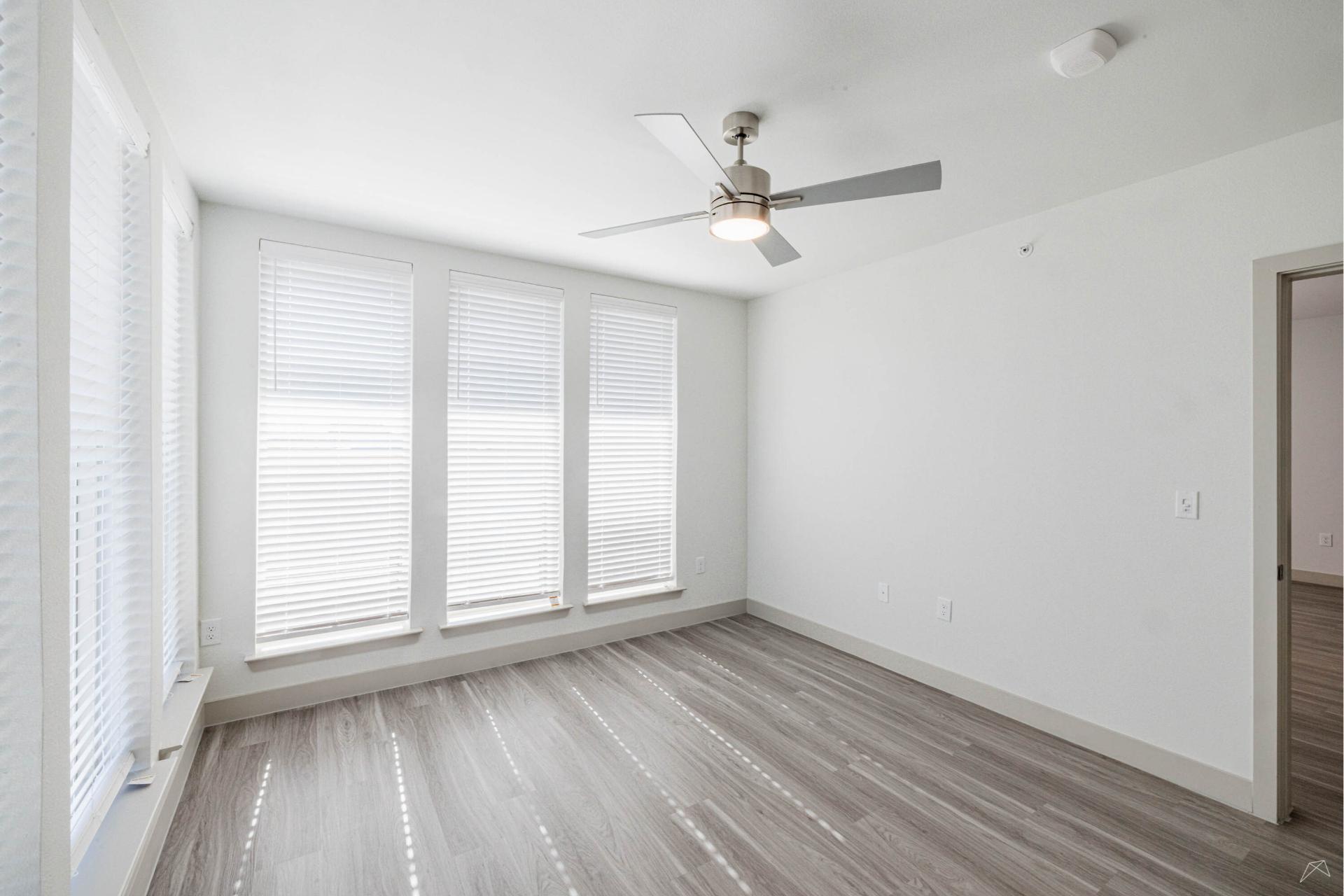 A bright, empty room with three tall windows covered by blinds, light gray wood-look flooring, white walls, a modern ceiling fan, and an open doorway leading to another room.