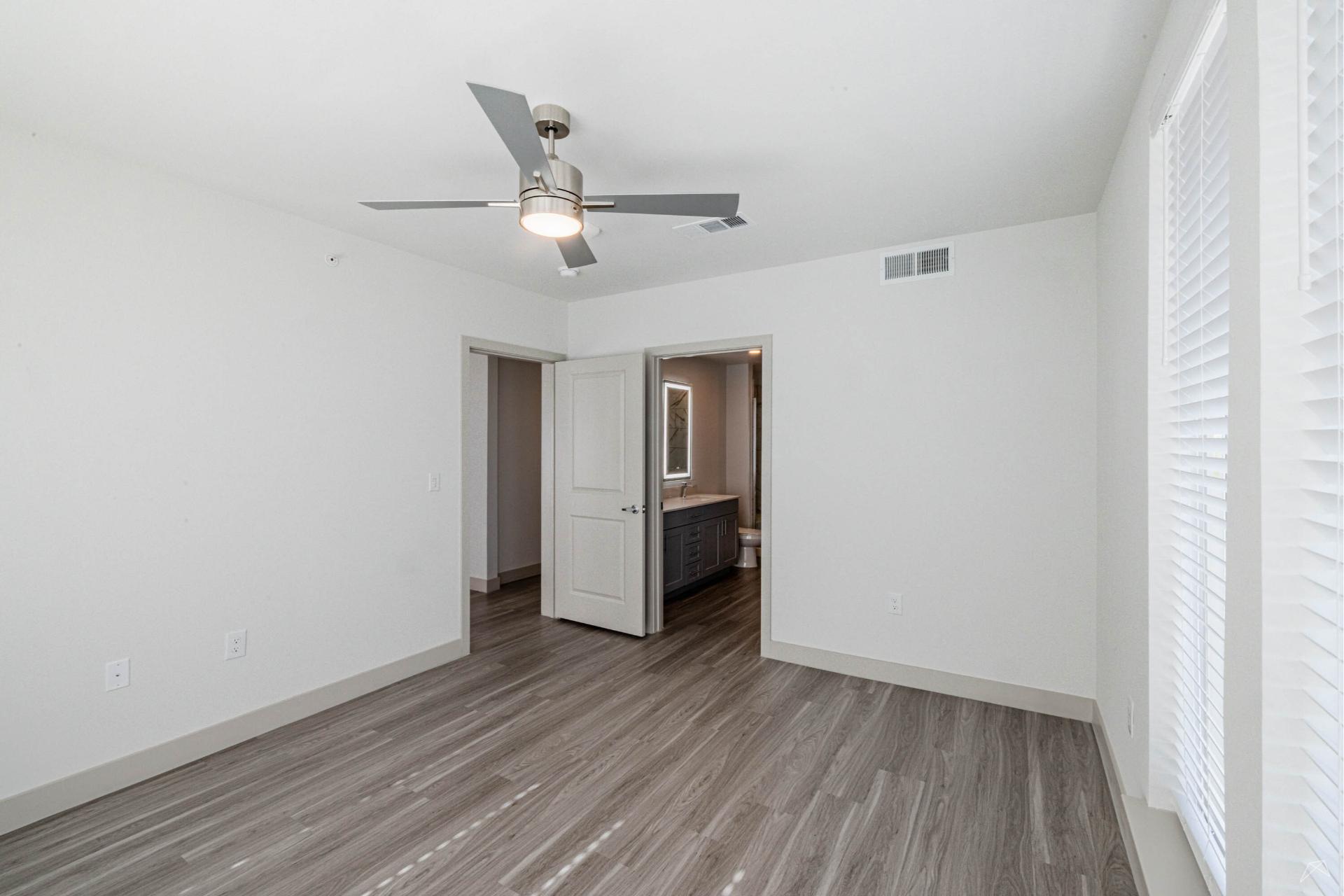 A bright, empty room with light gray wood-look flooring, white walls, ceiling fan, two open doors, and large windows letting in sunlight.
