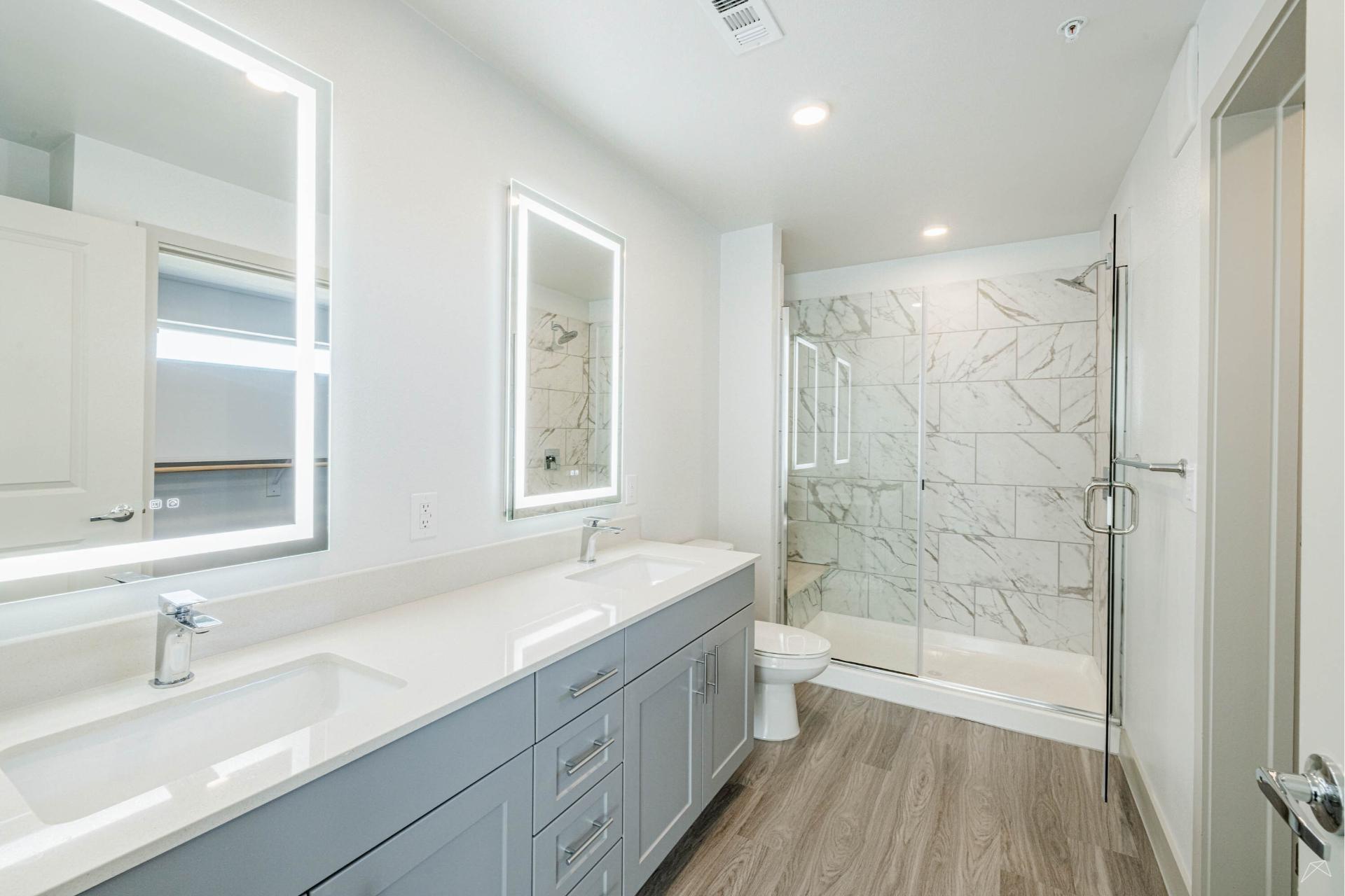 Modern bathroom with double sink vanity, lit mirrors, glass shower with marble tile, and wood-look flooring. Design is bright, minimalist.