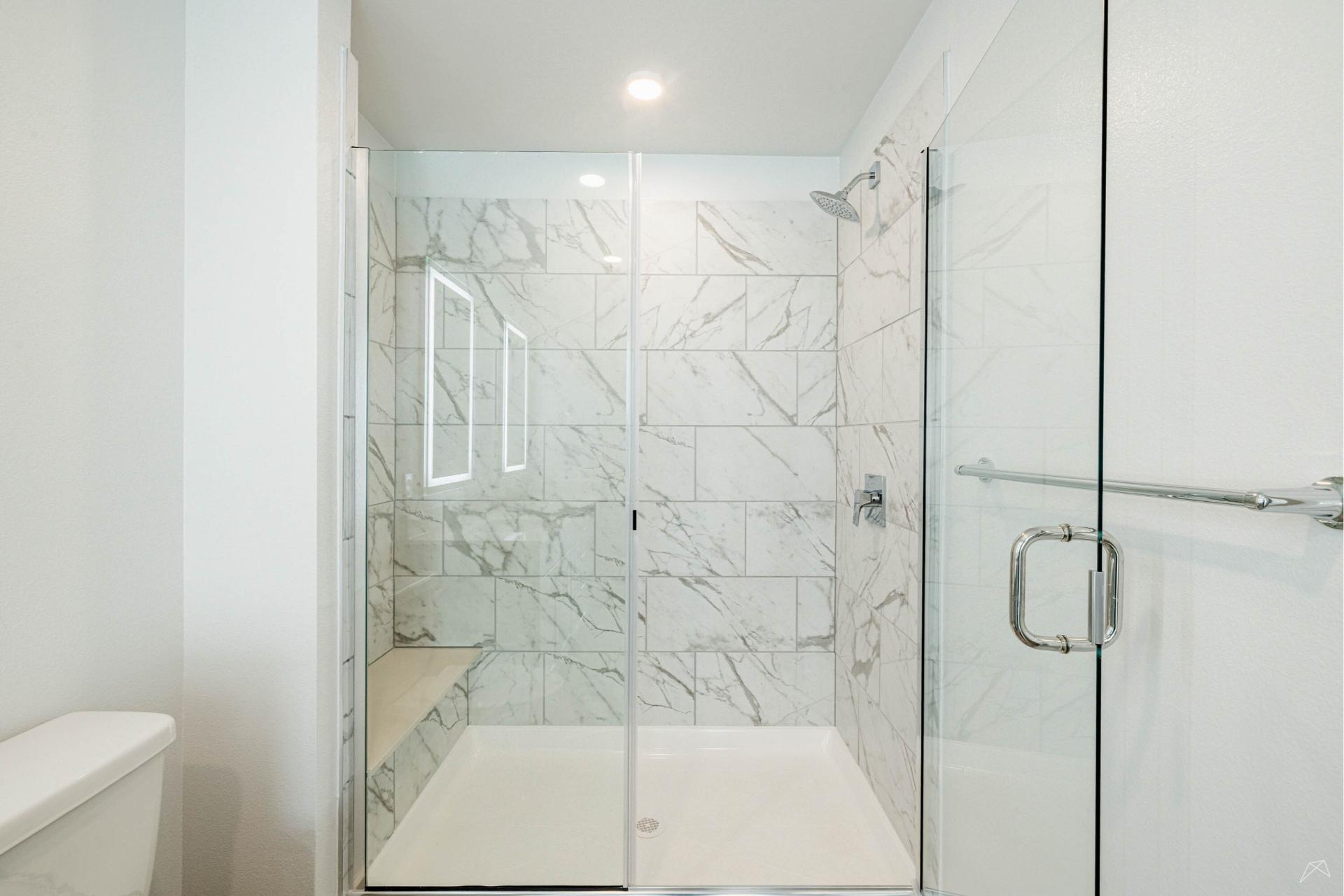 A modern shower with glass doors, white marble-patterned tiles, a built-in bench left, silver showerhead and handle right, toilet visible.