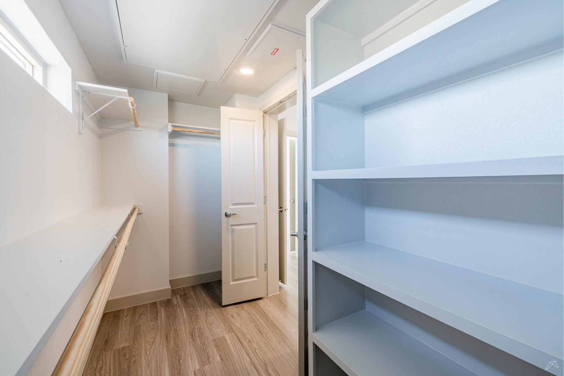 A spacious, empty walk-in closet with wood-look flooring, white shelves, a hanging rod, and a slightly open white door. Natural light enters.
