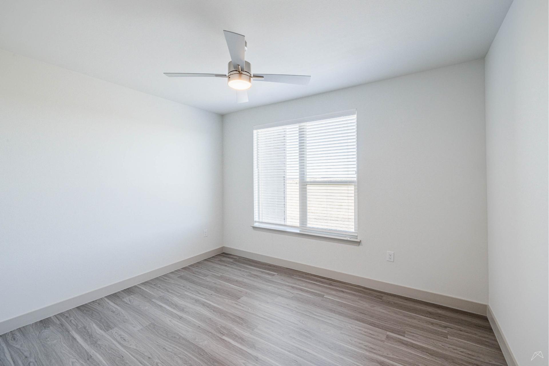 A bright, empty room with light gray wood-look flooring, white walls, a large window with blinds, and a modern ceiling fan with a light.