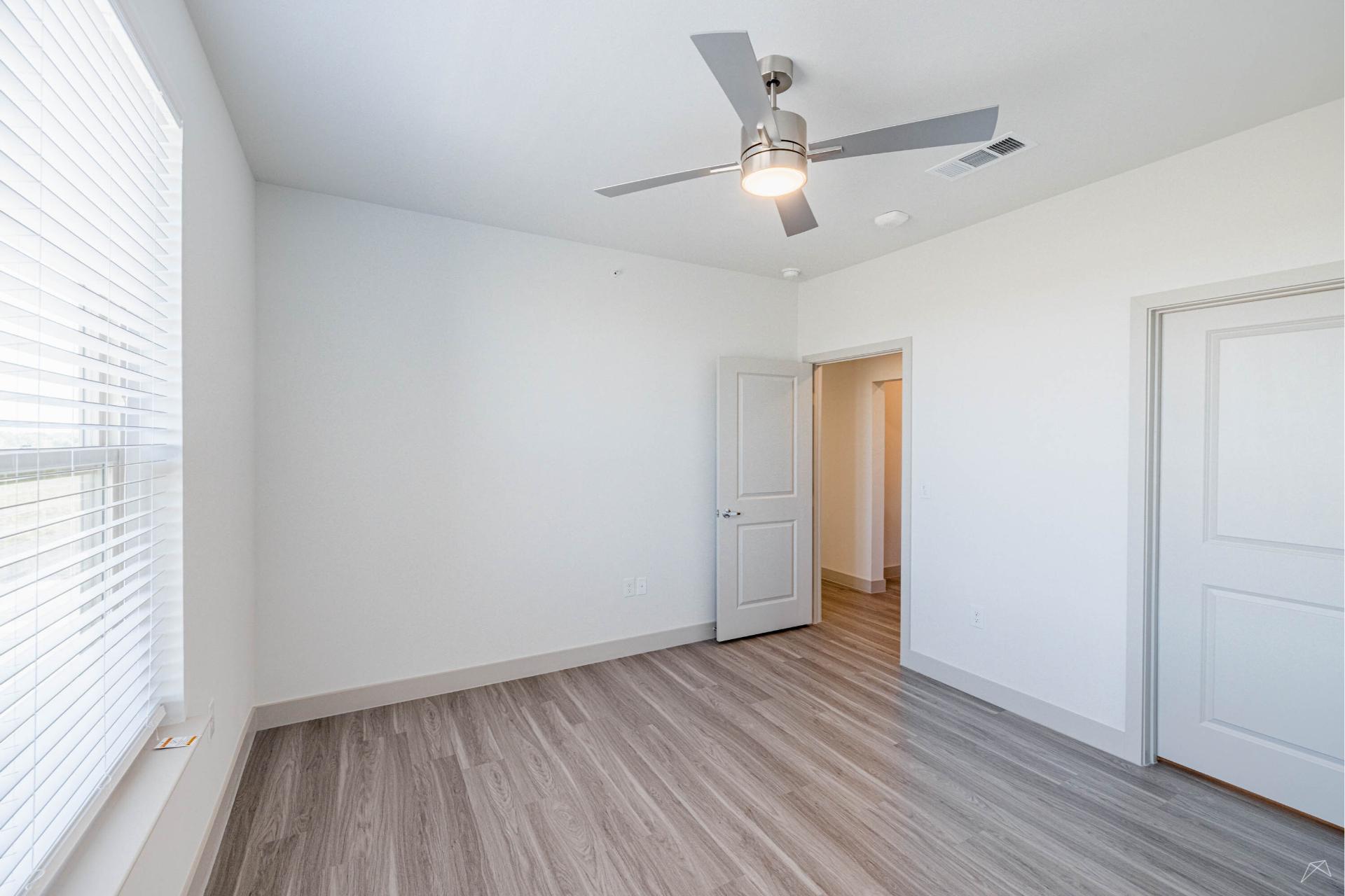A bright, empty room with light wood-look flooring, white walls, a ceiling fan with a light, two closed doors, and a sunny window.
