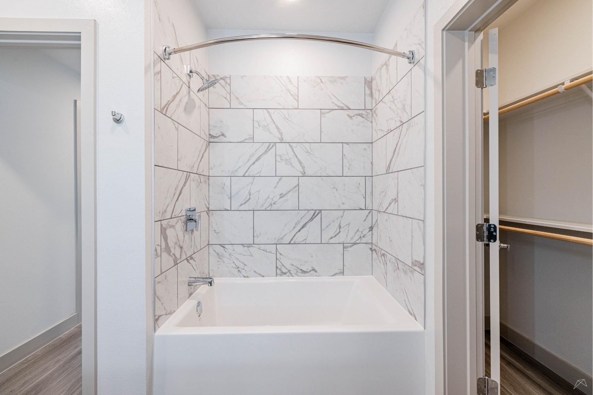 A modern bathroom with a white bathtub, marble-patterned tile walls, curved shower rod, and a closet with wood-like shelves and rods.