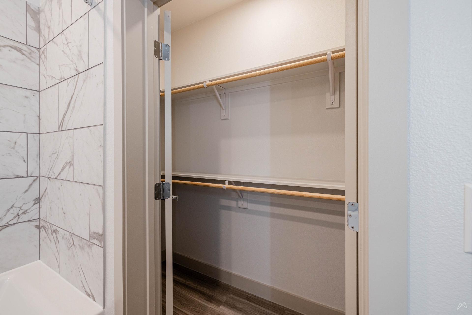 A walk-in closet with double hanging wood-like rods and empty shelves, beside a bathroom with white marble-patterned tile walls and a bathtub.