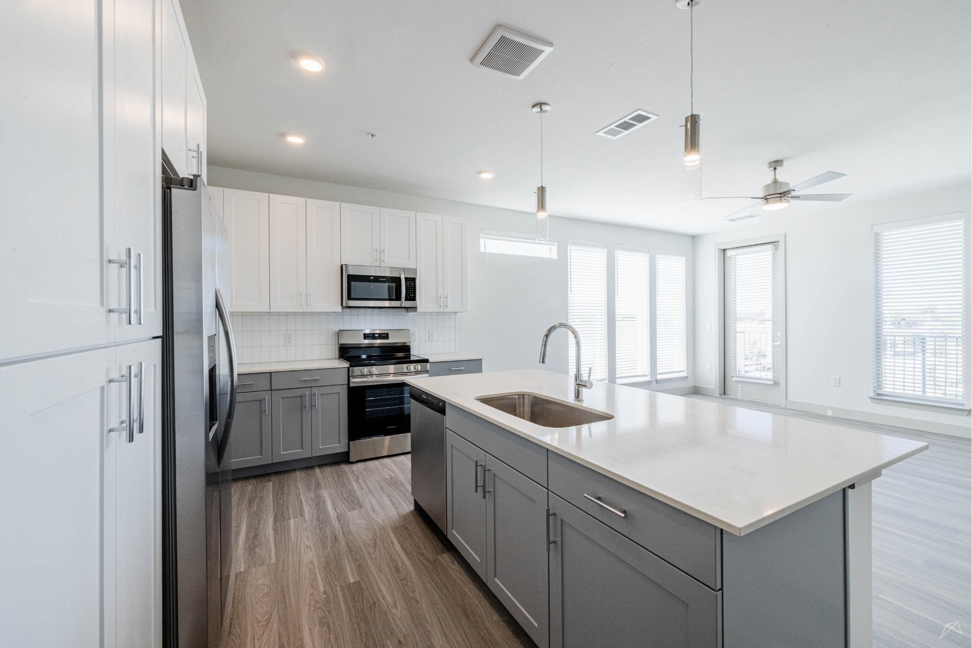 Modern kitchen with gray lower, white upper cabinets, large island with sink, stainless appliances, wood-look flooring, open living area.