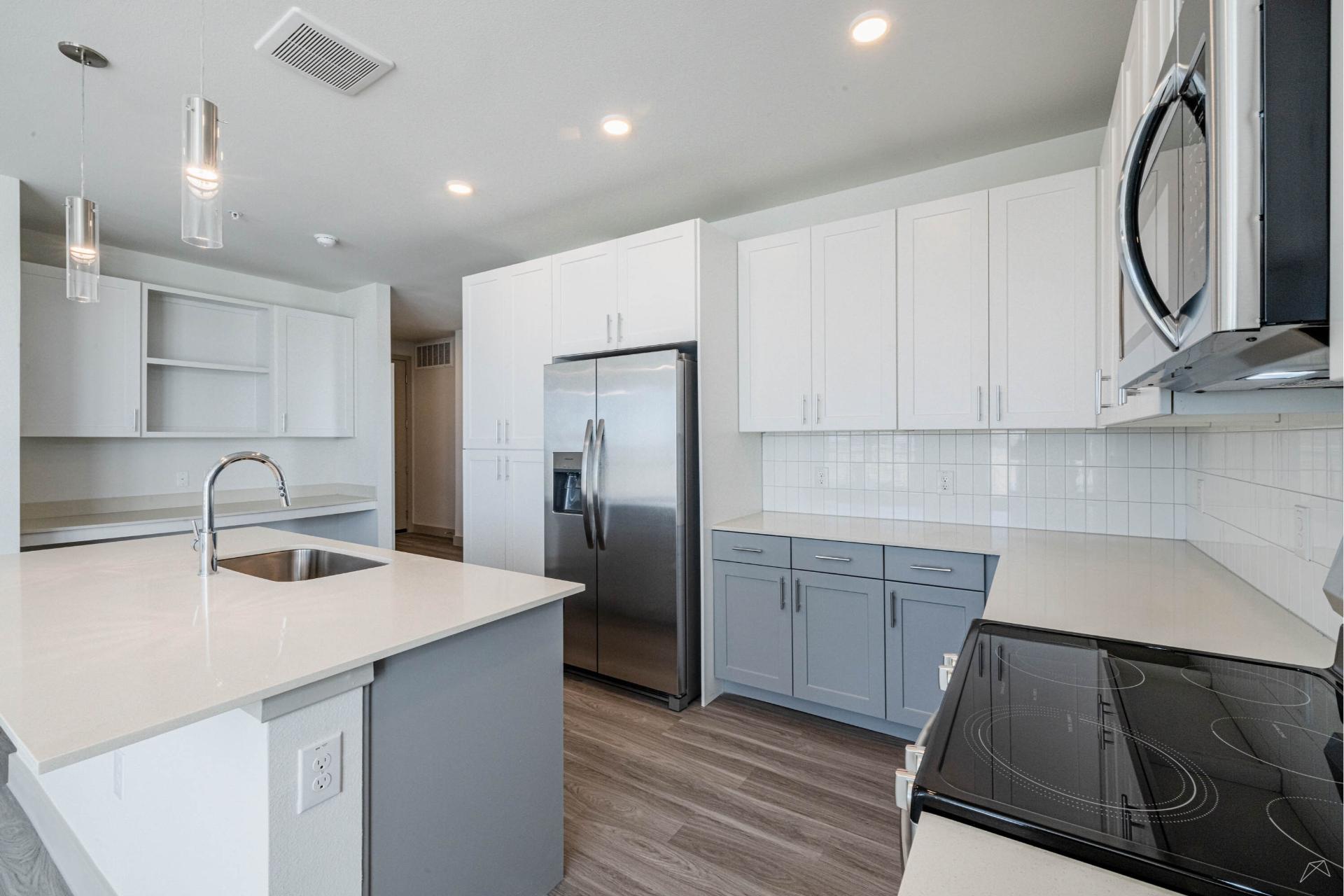 Modern kitchen with stainless steel appliances, white upper cabinets, gray lowers, white countertops, island sink, wood-look flooring.