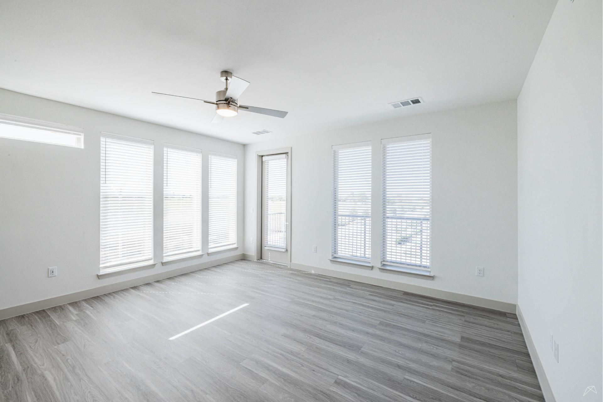 A bright, empty room with light gray wood-look flooring, white walls, big windows with blinds, a ceiling fan, and a door outside.