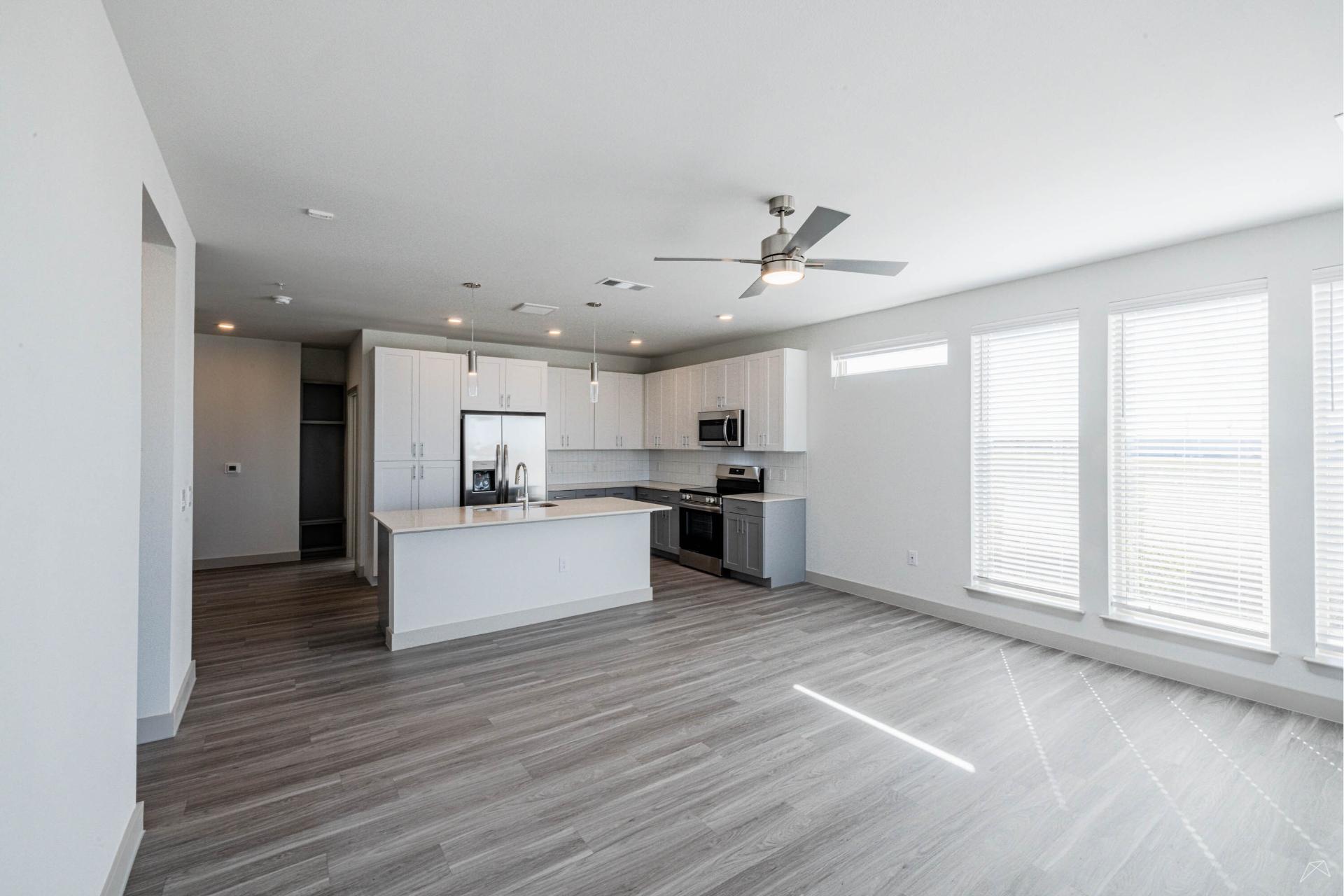 A modern, unfurnished apartment with light gray wood-like flooring, white walls, large windows, a ceiling fan, and an open kitchen.