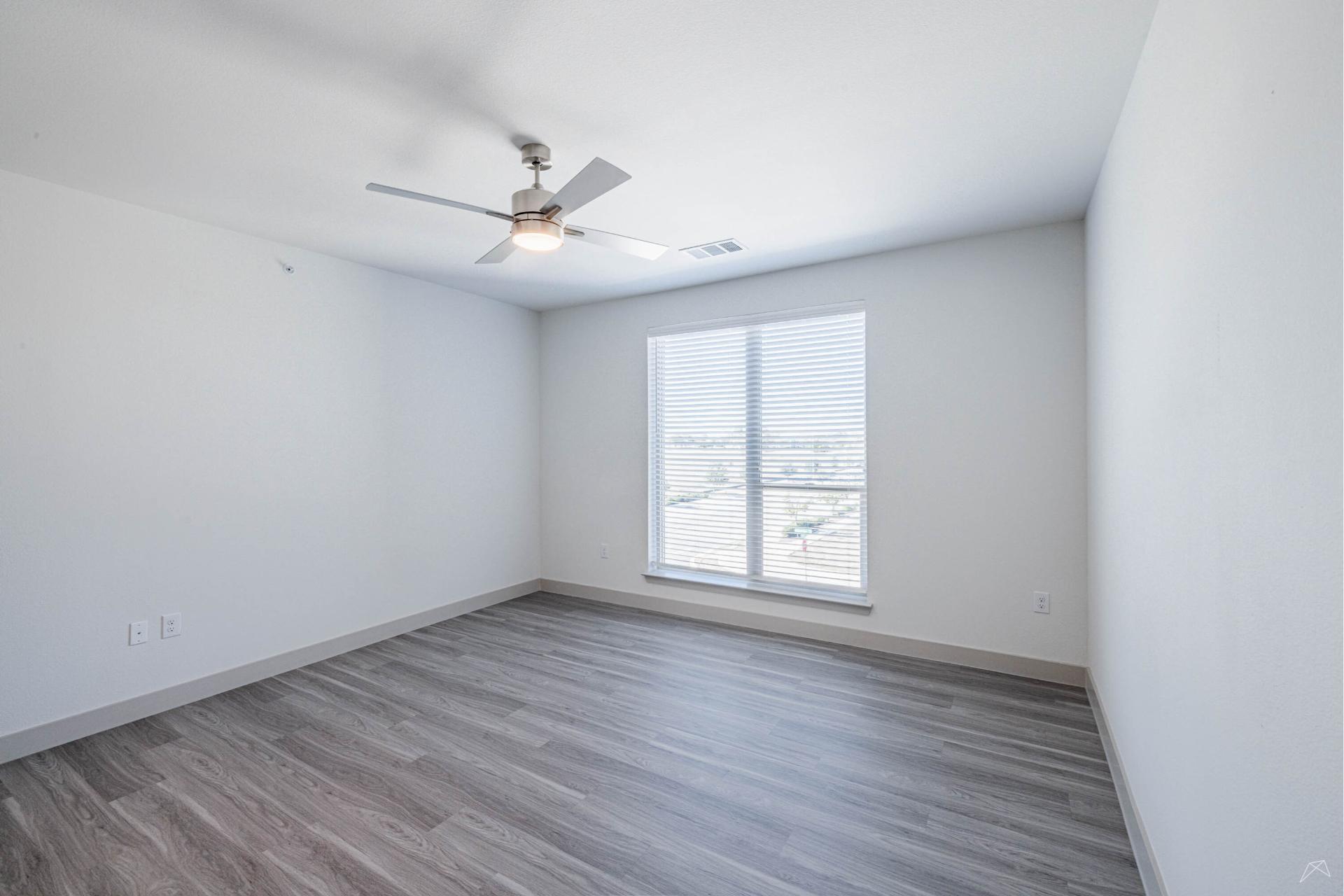 A bright, empty room with light gray walls, wood-look flooring, a ceiling fan with a light, and a large window with blinds letting in sunlight.