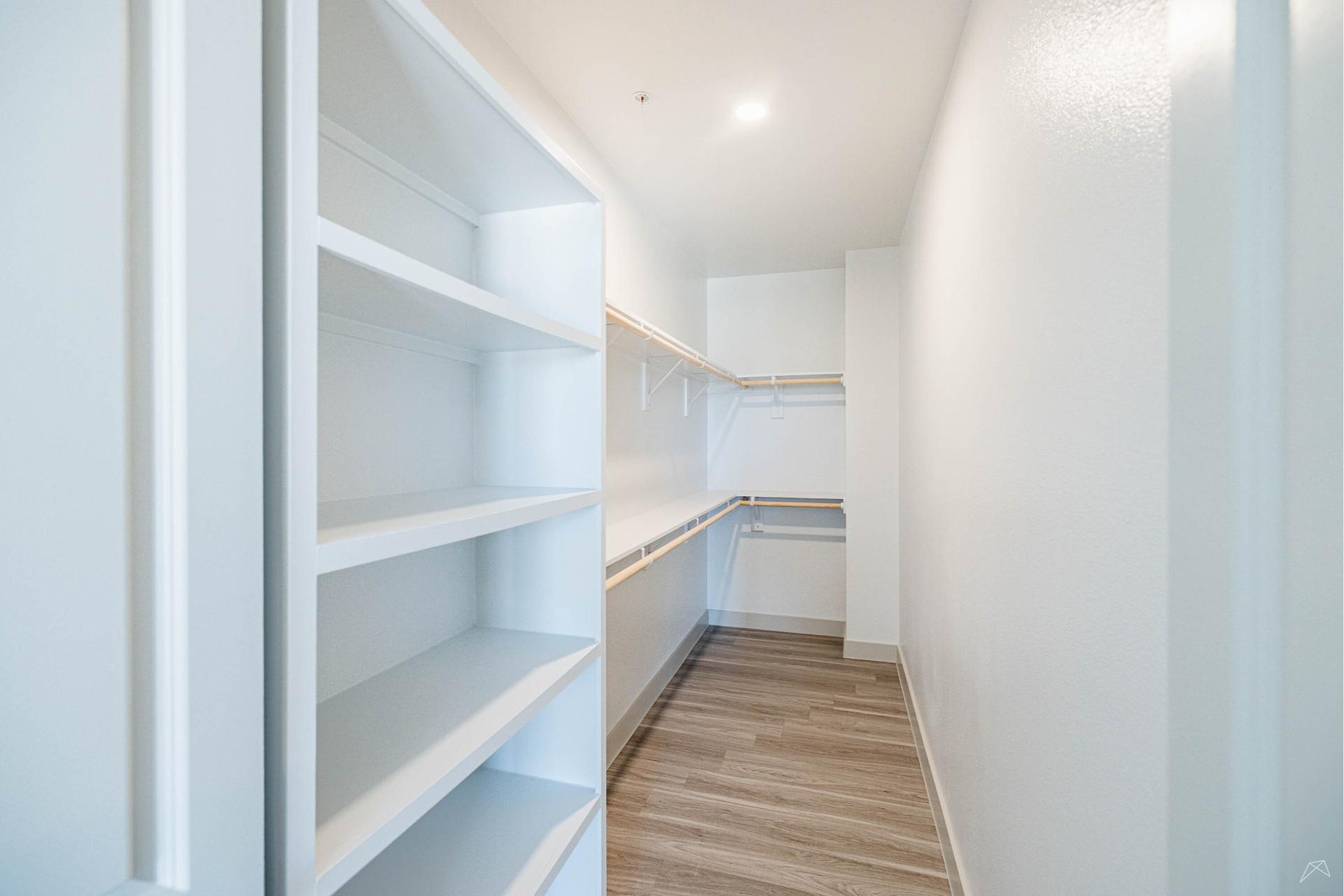 A spacious, empty walk-in closet with built-in white shelves, hanging rods on both sides, light walls, and wood-look flooring.