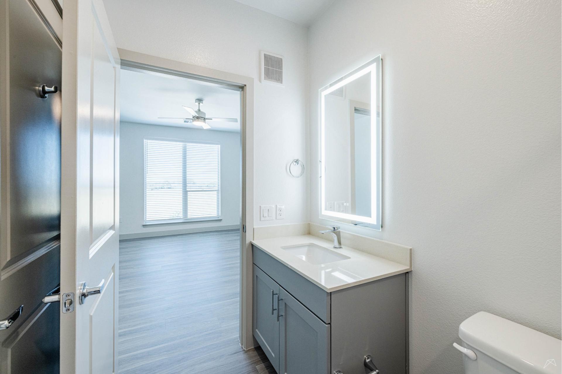 A modern bathroom with a gray vanity, lighted mirror, and white walls, next to a bright bedroom with large windows and wood-like flooring.