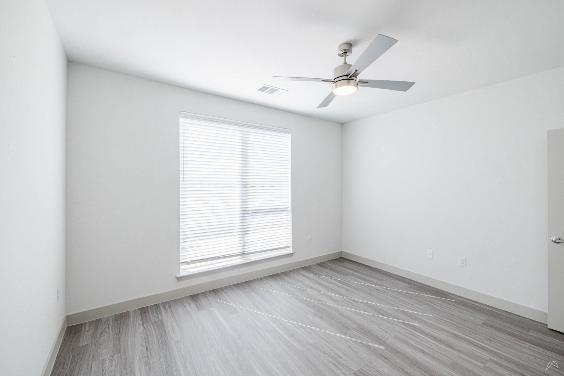 A bright, empty room with white walls, a large window with blinds, gray wood-look flooring, and a ceiling fan with light fixture.
