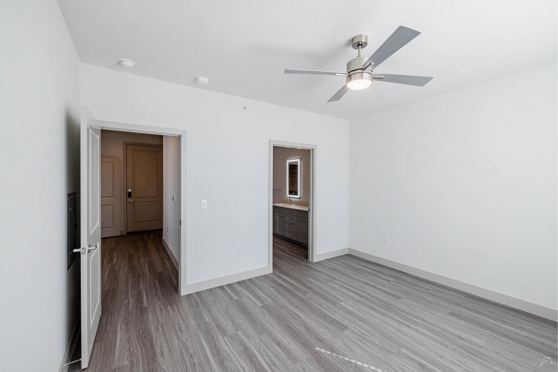 A modern empty bedroom with light gray wood-look flooring, white walls, a ceiling fan with light, and two doorways to hall and bathroom.