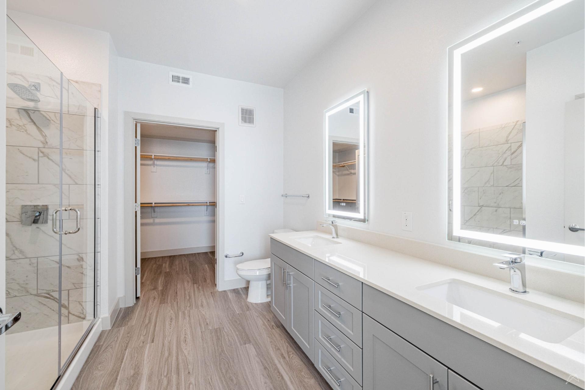 Modern bathroom with light wood-look flooring, double-sink vanity with gray cabinets, large mirrors, glass shower, and open walk-in closet.
