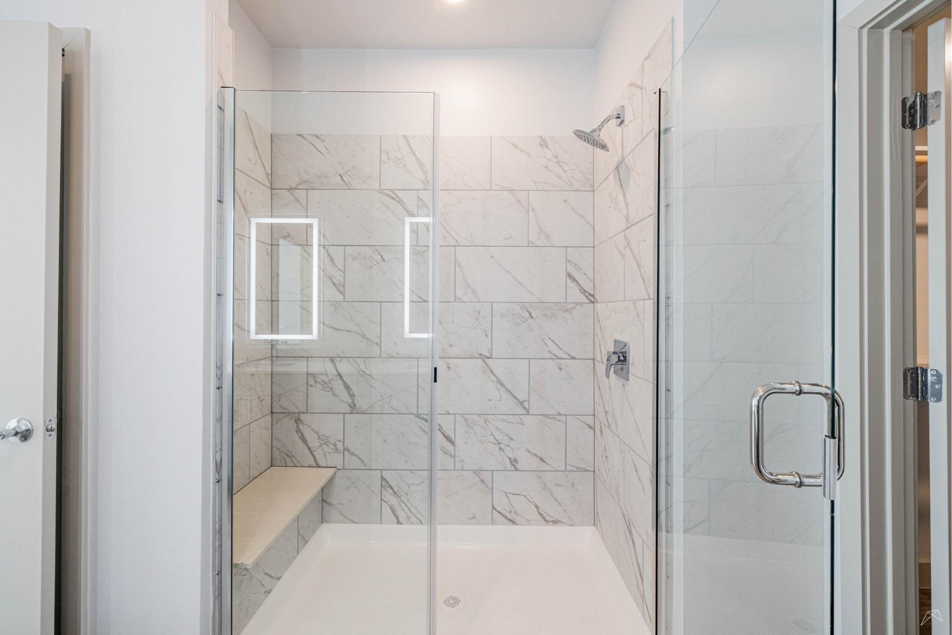 A modern bathroom shower with glass doors, marble-patterned tile walls, a wood-look built-in bench, and a wall-mounted showerhead.
