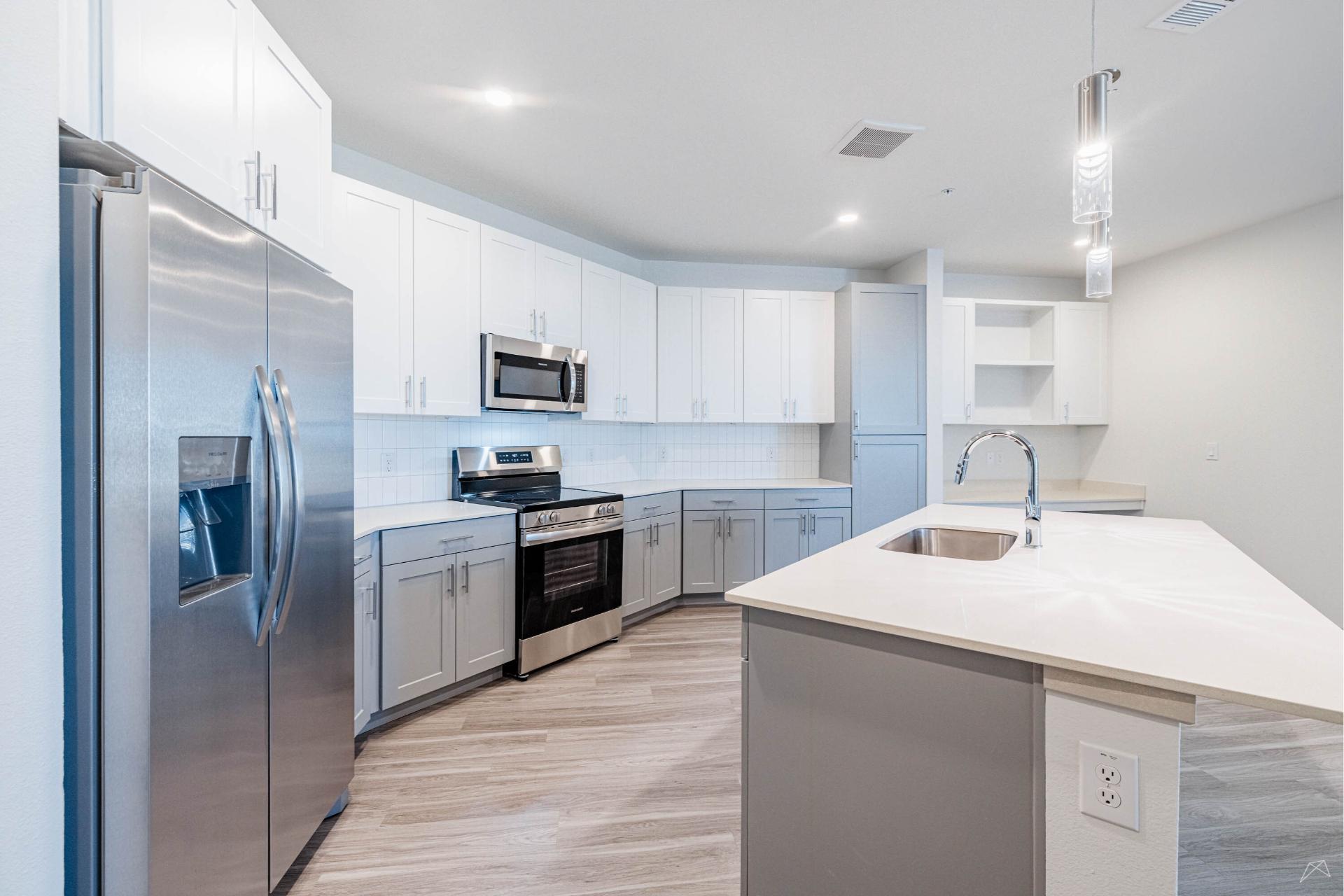 Modern kitchen with white and gray cabinets, stainless appliances, large fridge, island sink, wood-look flooring, and recessed lighting.