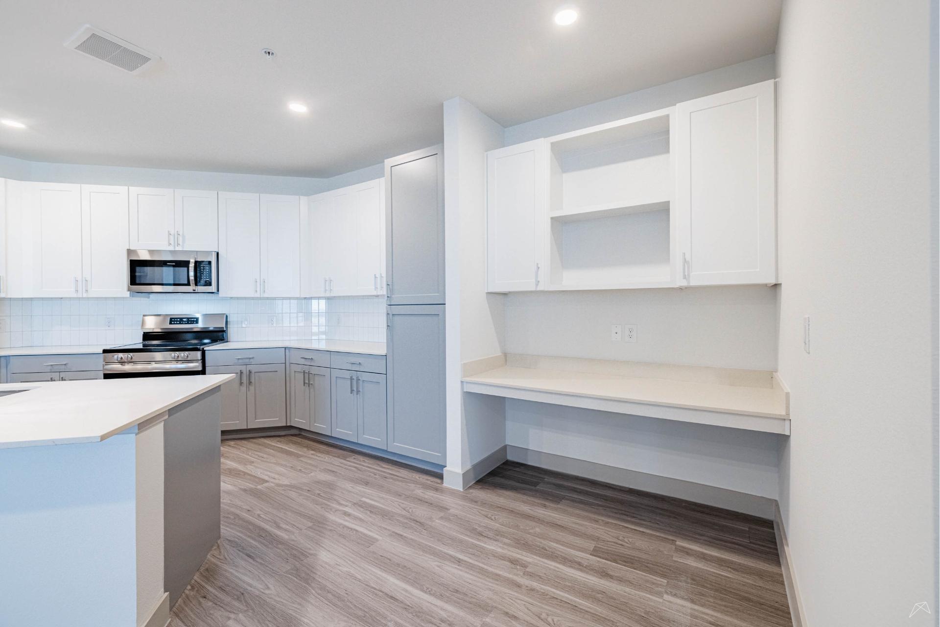 Modern kitchen with white and gray cabinets, stainless steel appliances, wood-look flooring, and built-in desk area; bright and minimalist.