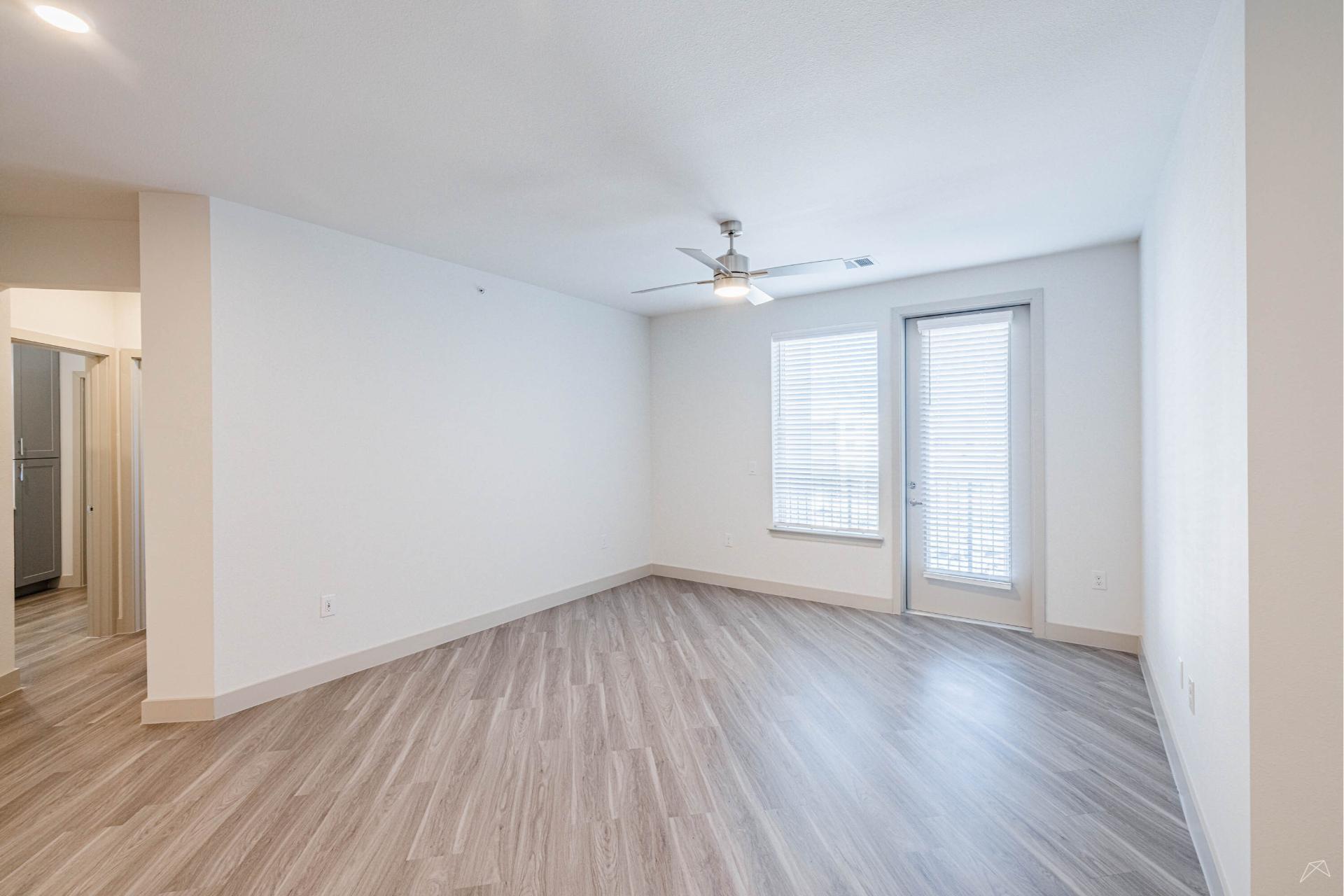 A bright, empty room with light wood-look flooring, white walls, a ceiling fan, large windows with blinds, and a glass door outside.