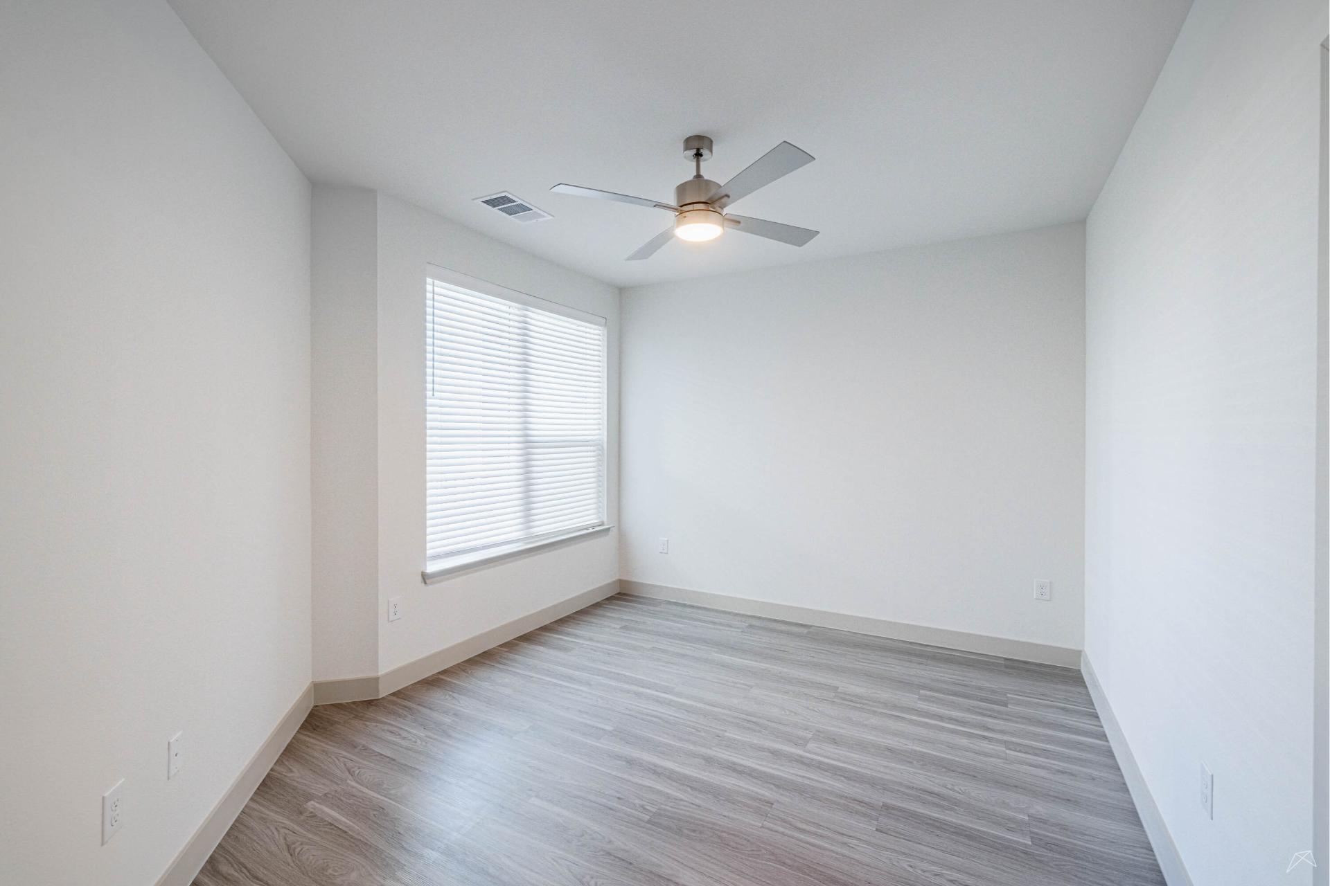 A bright, empty room with light gray wood-look flooring, white walls, a large window with blinds, and a ceiling fan with light fixture.
