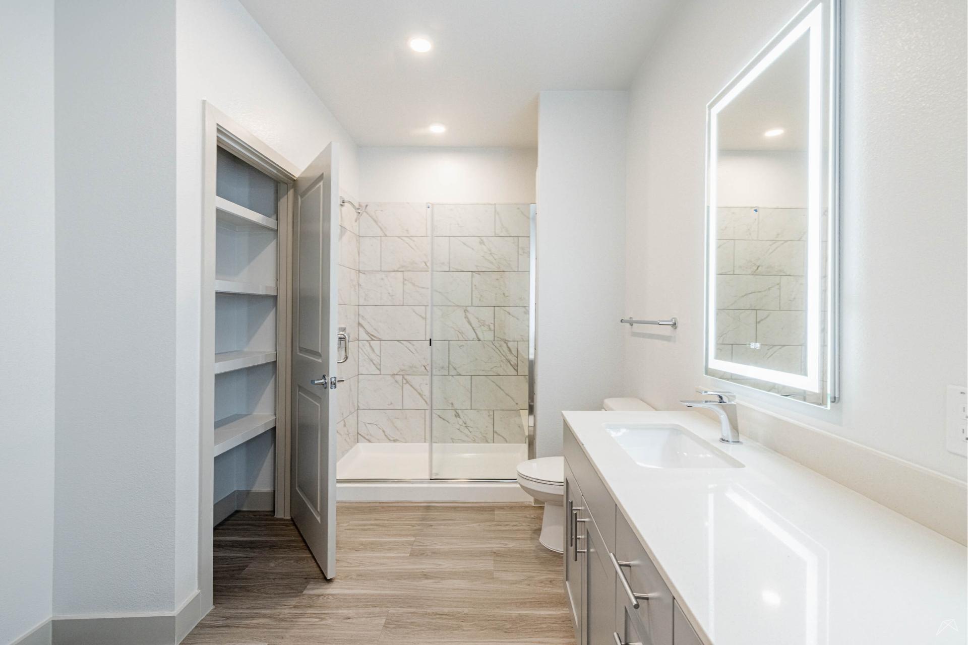 Modern bathroom with light wood-look flooring, white walls, built-in open shelves, a large vanity, illuminated mirror, and glass shower.