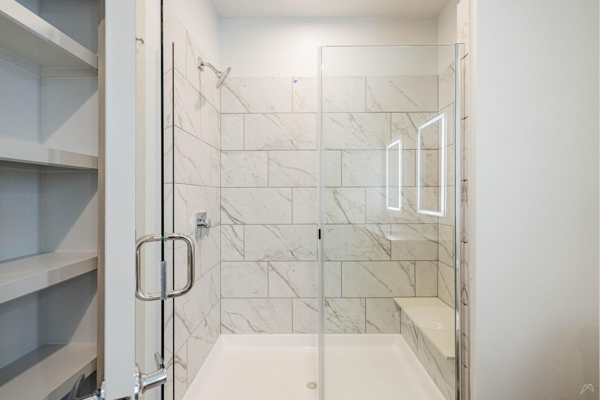 Modern shower with white and gray marble tiles, glass door, built-in bench on the right, and wood-look open shelving on the left.