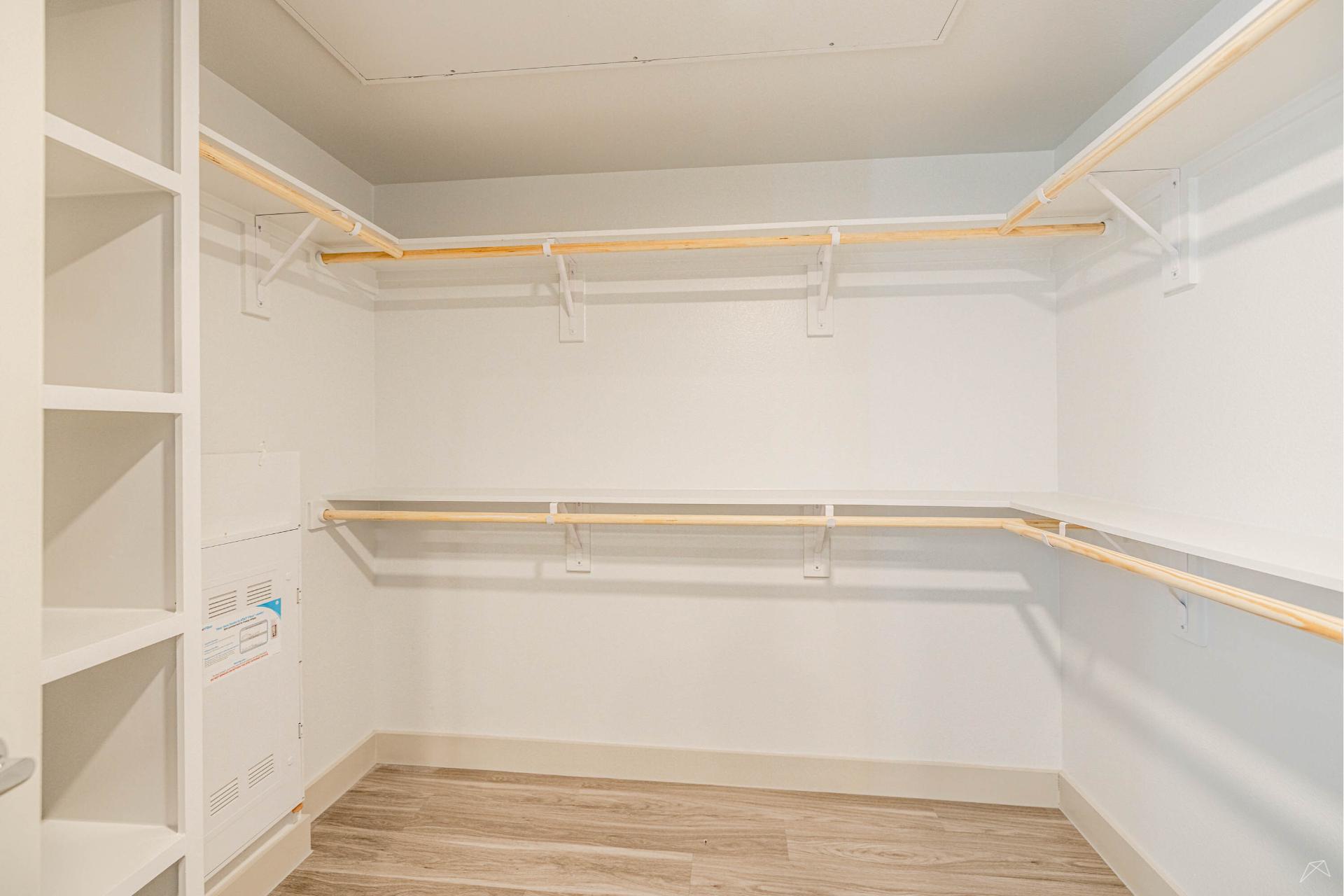 Empty walk-in closet with light-colored walls, wood-like shelves, open hanging rods on two sides, laminate floor, and a white utility panel.