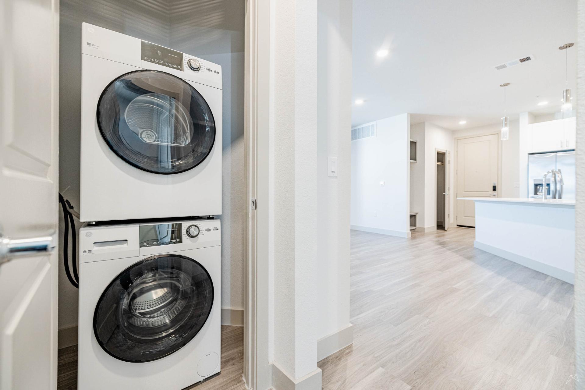 A modern apartment with stacked washer and dryer in a white-walled nook, wood-look flooring, next to an open kitchen and hallway.