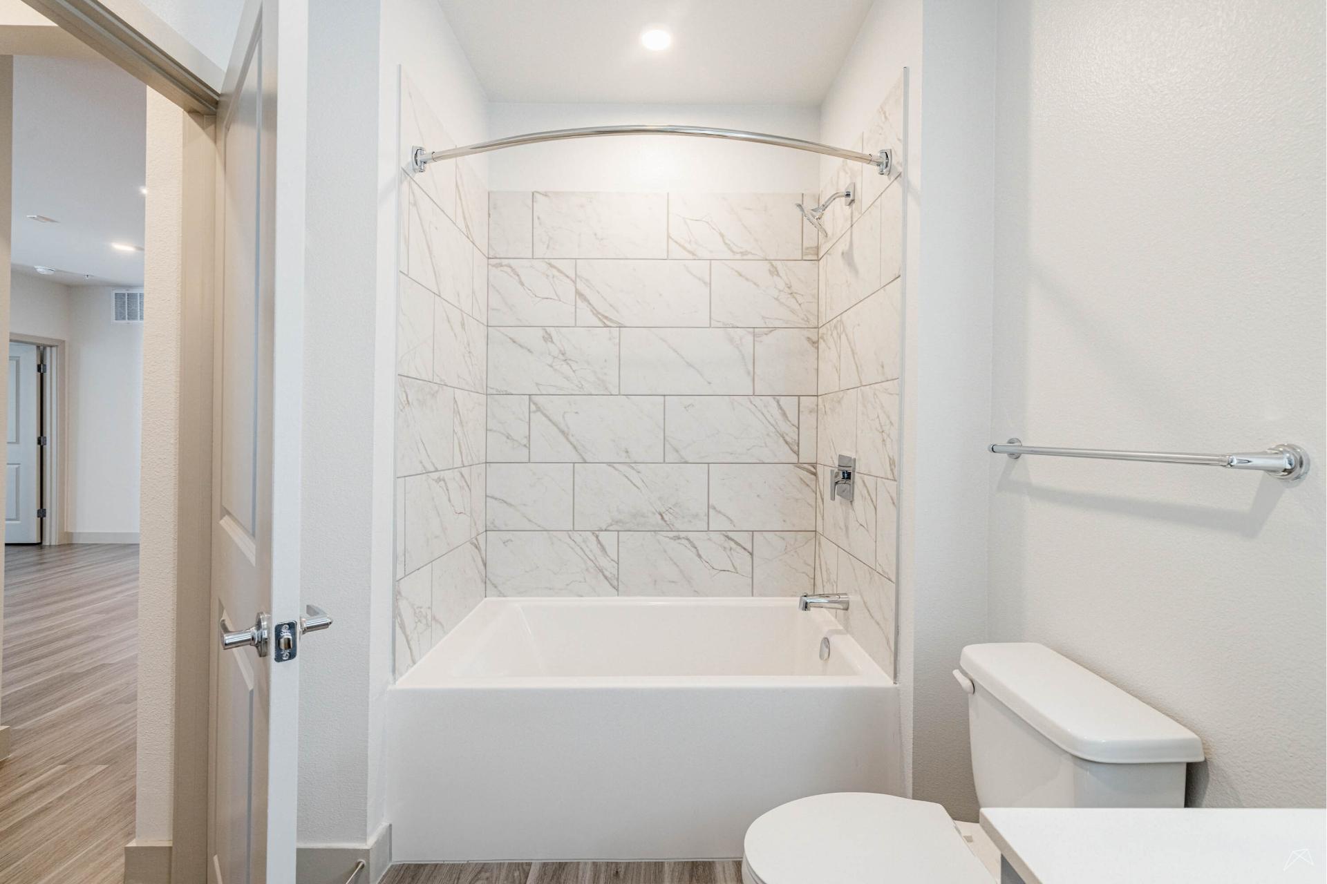 Modern bathroom with a white bathtub and shower, marble-pattern tiled walls, toilet, towel bar, wood-look flooring, bright white walls, and recessed lighting.