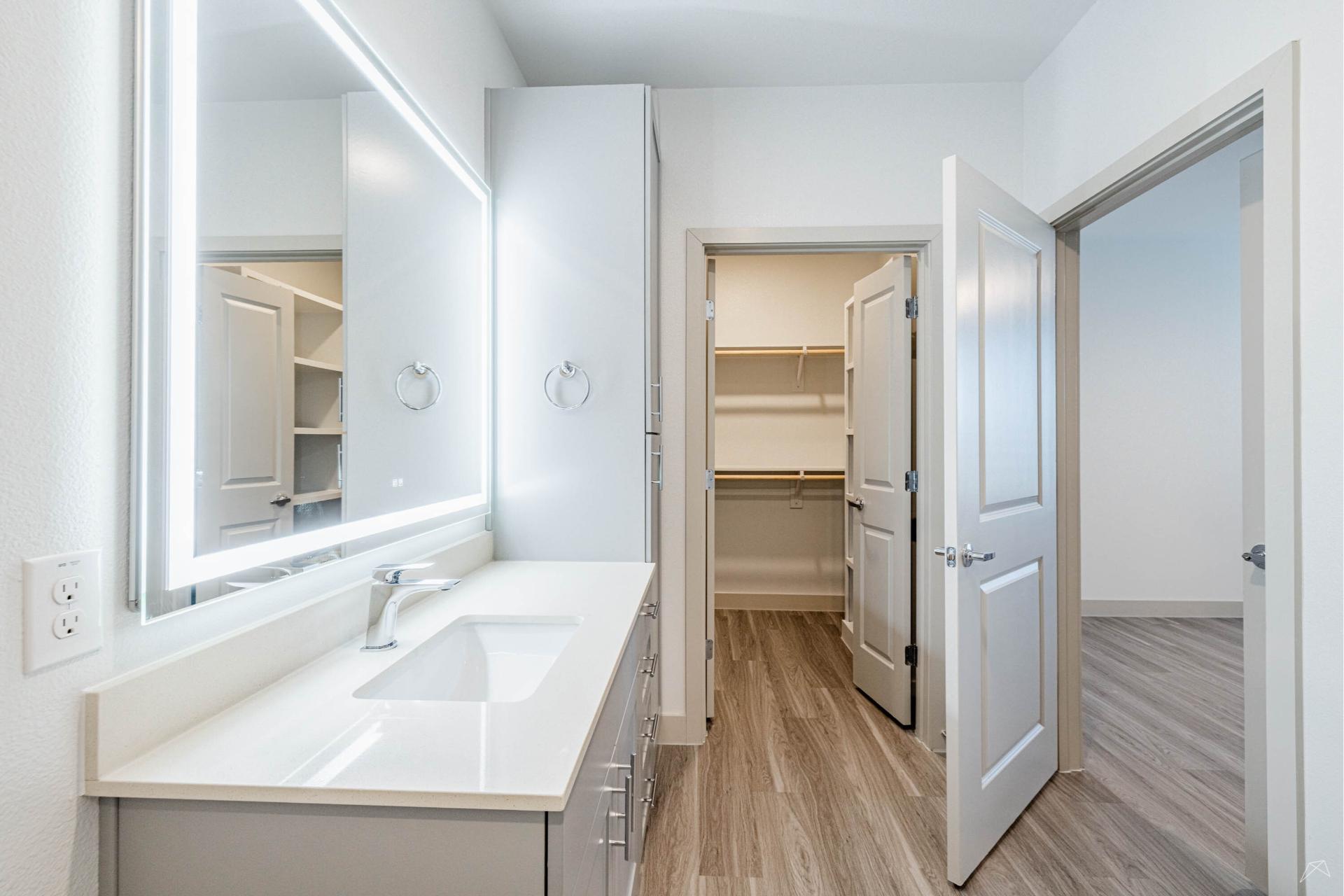 Modern bathroom with light wood-look flooring, large illuminated mirror above a white sink and countertop, open door to walk-in closet.