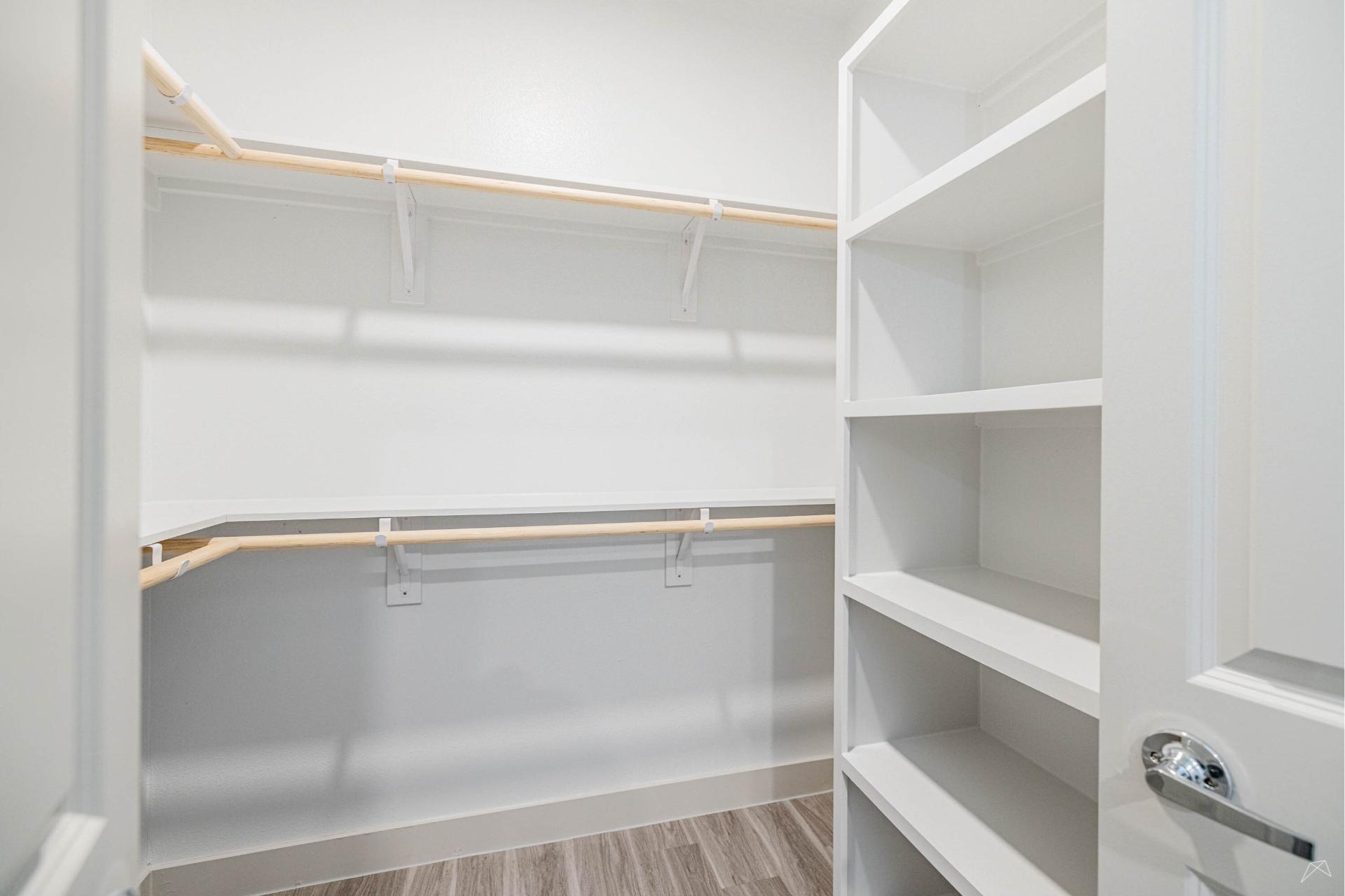 A clean, empty walk-in closet with white shelves, wood-style hanging rods, light wood-style flooring, white walls, and an open door.