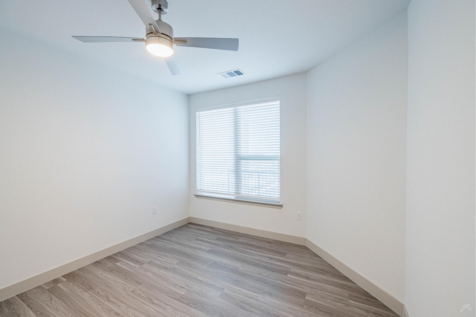 Empty room with light gray wood-style flooring, white walls, ceiling fan with a light, and large window with closed blinds letting in daylight.