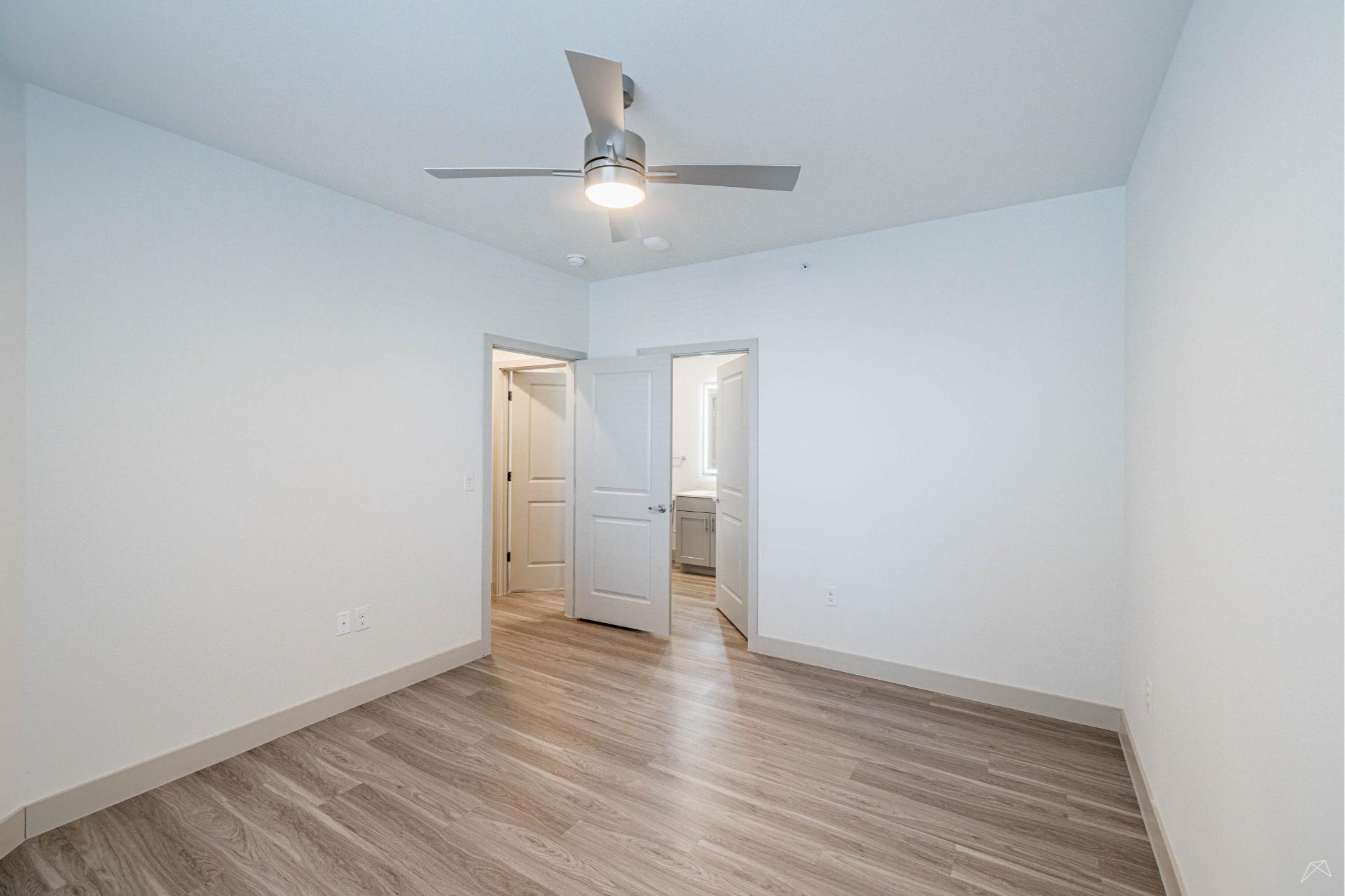 Empty, unfurnished room with light-colored walls, wood-like flooring, ceiling fan with light, and open door to a bright hallway and bathroom.