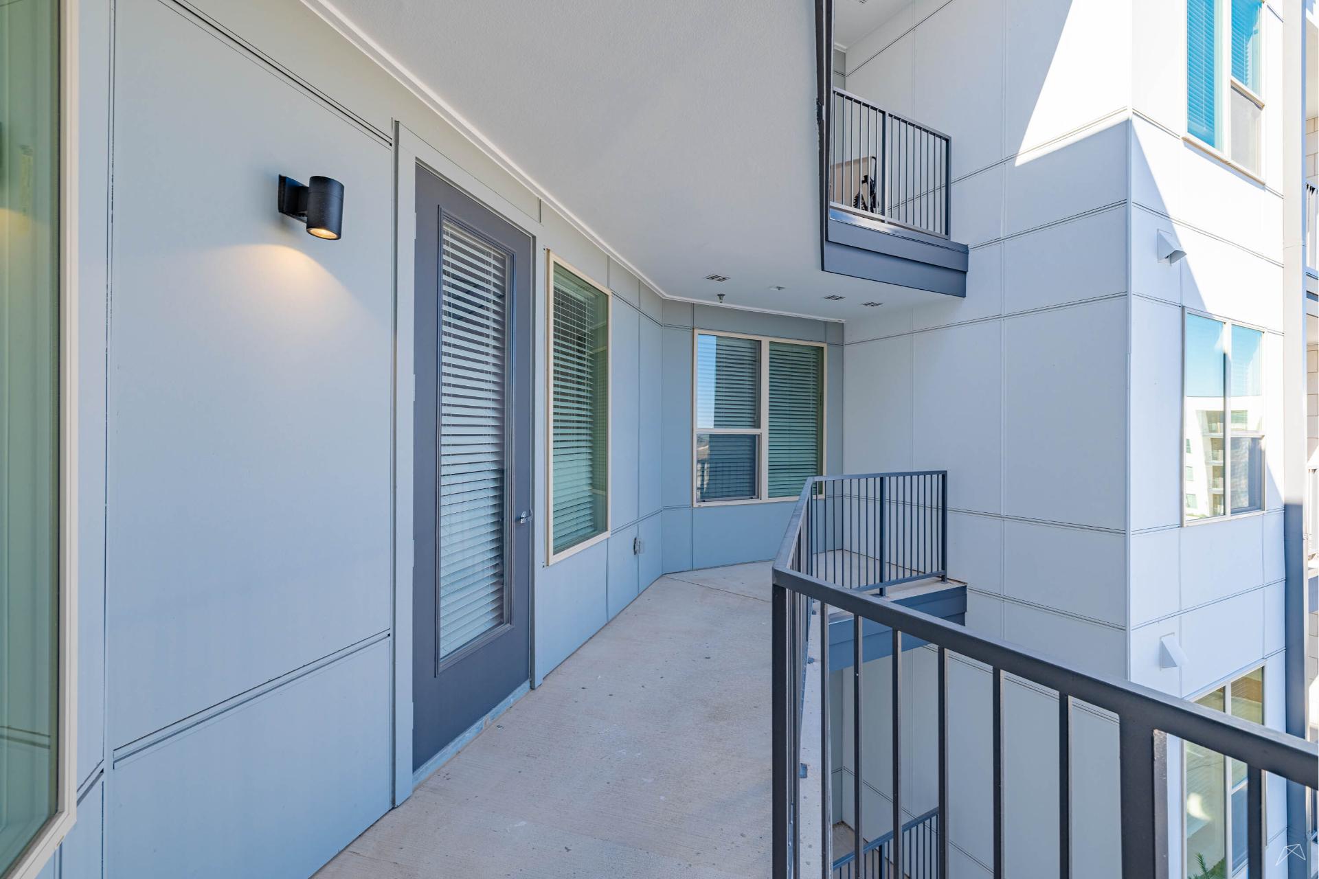 A modern apartment balcony with light blue walls, metal railings, a wood-look door with blinds, large windows, and a concrete floor.